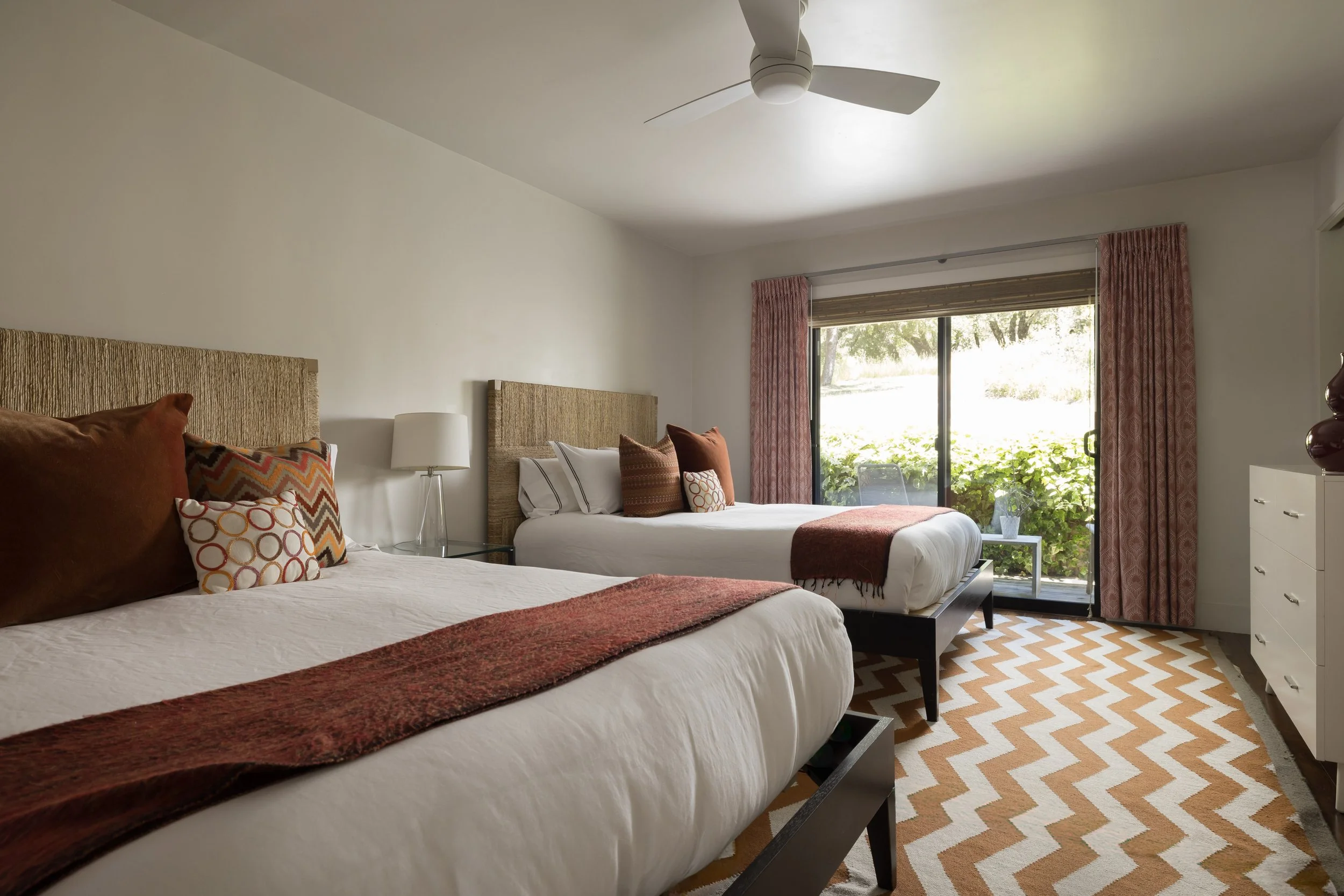 Two beds with white linens and decorative pillows, a sliding glass door with pink curtains leading to an outdoor patio, and a chevron patterned area rug in the room.