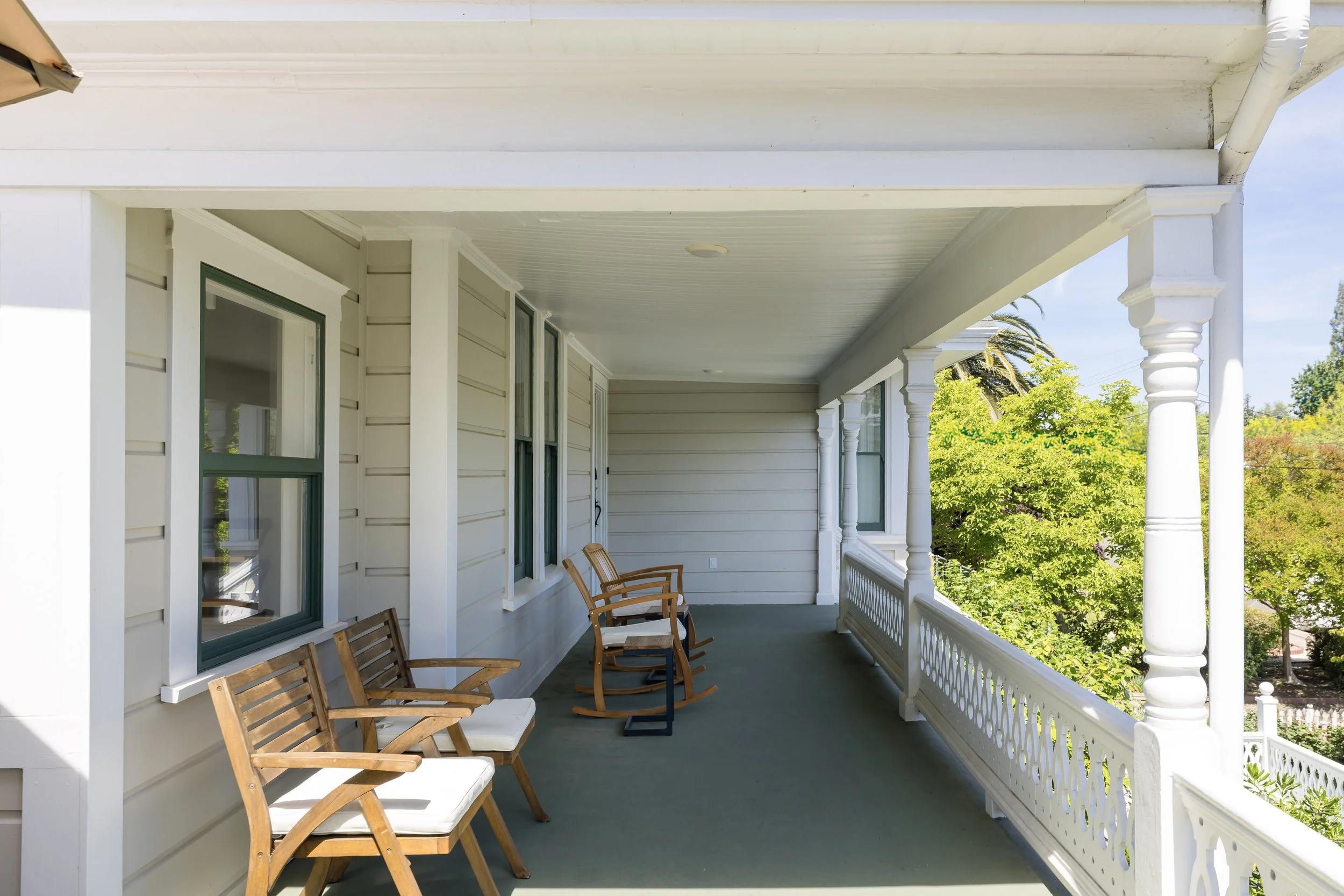A spacious porch with white siding, wooden chairs, and a decorative white railing, surrounded by green trees and blue sky.