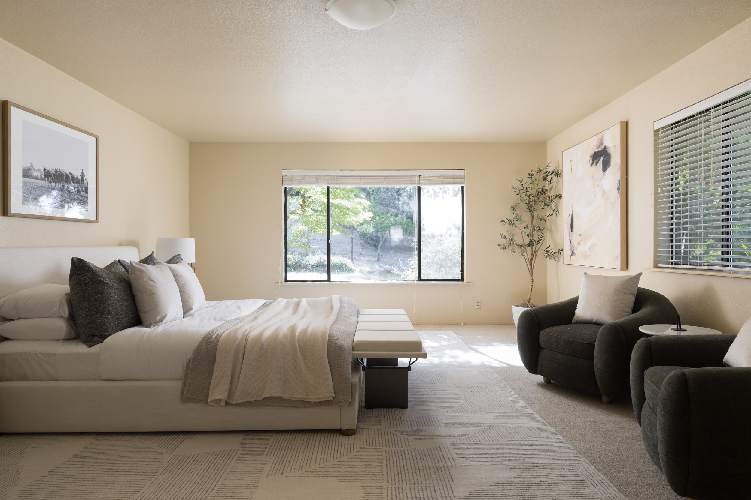 Living room with a white couch with gray and white pillows, black side table, gray armchair with a beige pillow, large window with blinds letting in natural light, artwork on beige walls, and a potted plant.