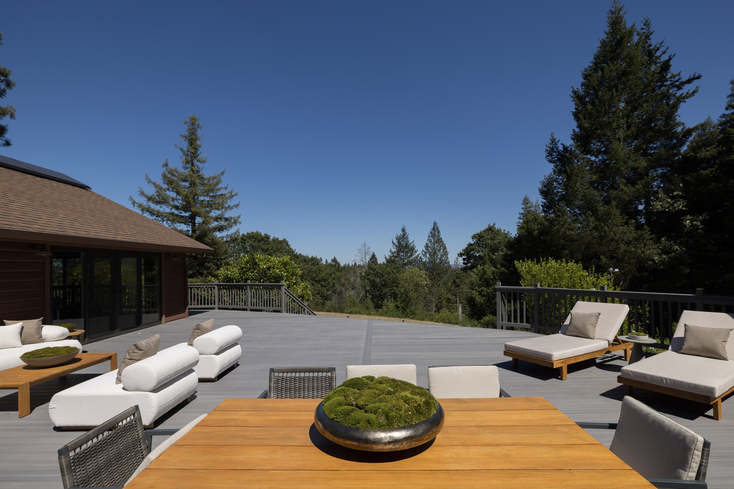 A spacious outdoor deck with white cushioned lounge chairs, a wooden dining table with a decorative moss bowl, and a view of lush green trees under a clear blue sky.