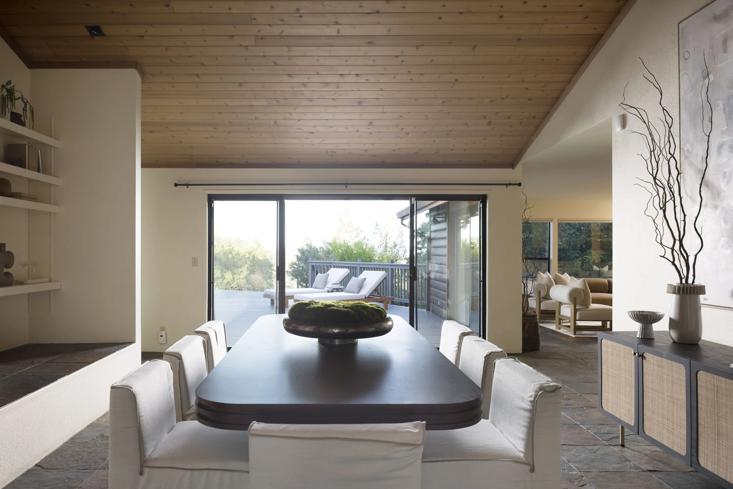 Interior of a modern dining room with a large rectangular table surrounded by white upholstered chairs, a decorative black bowl centerpiece, and a view of a porch with outdoor seating through glass sliding doors, with a wooden ceiling and minimalist 