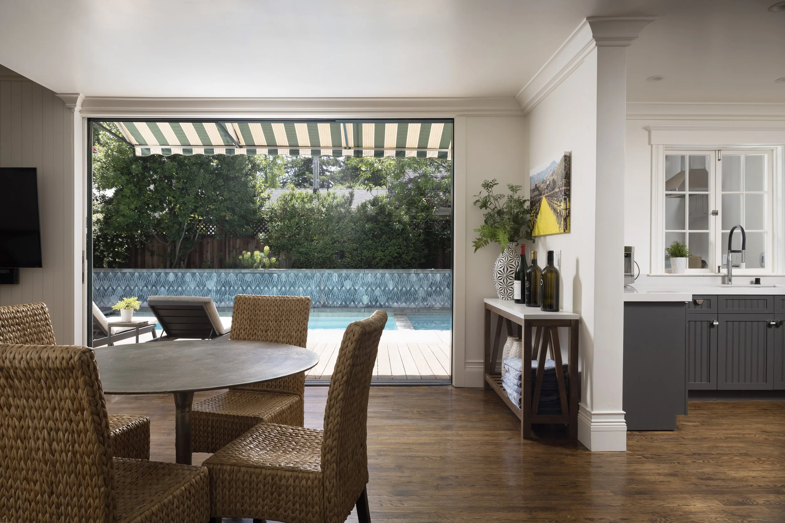 Interior view of a living and dining area with a sliding glass door leading to a backyard with pool, trees, and patio furniture. The space features wicker chairs, a round wooden table, a side table with decorative vases, plants, artwork, and a kitche