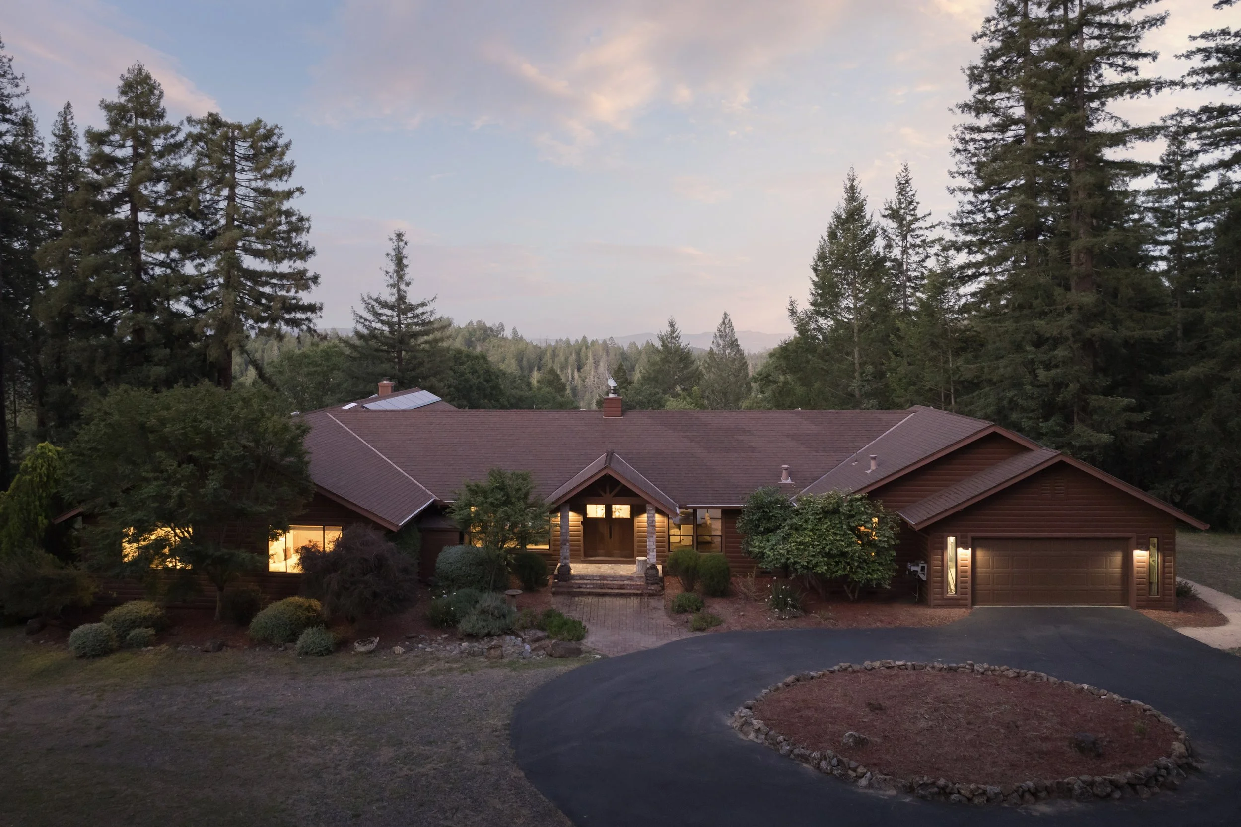 A house with a red roof and large windows, surrounded by trees and a driveway, at dusk or dawn in a forested area.