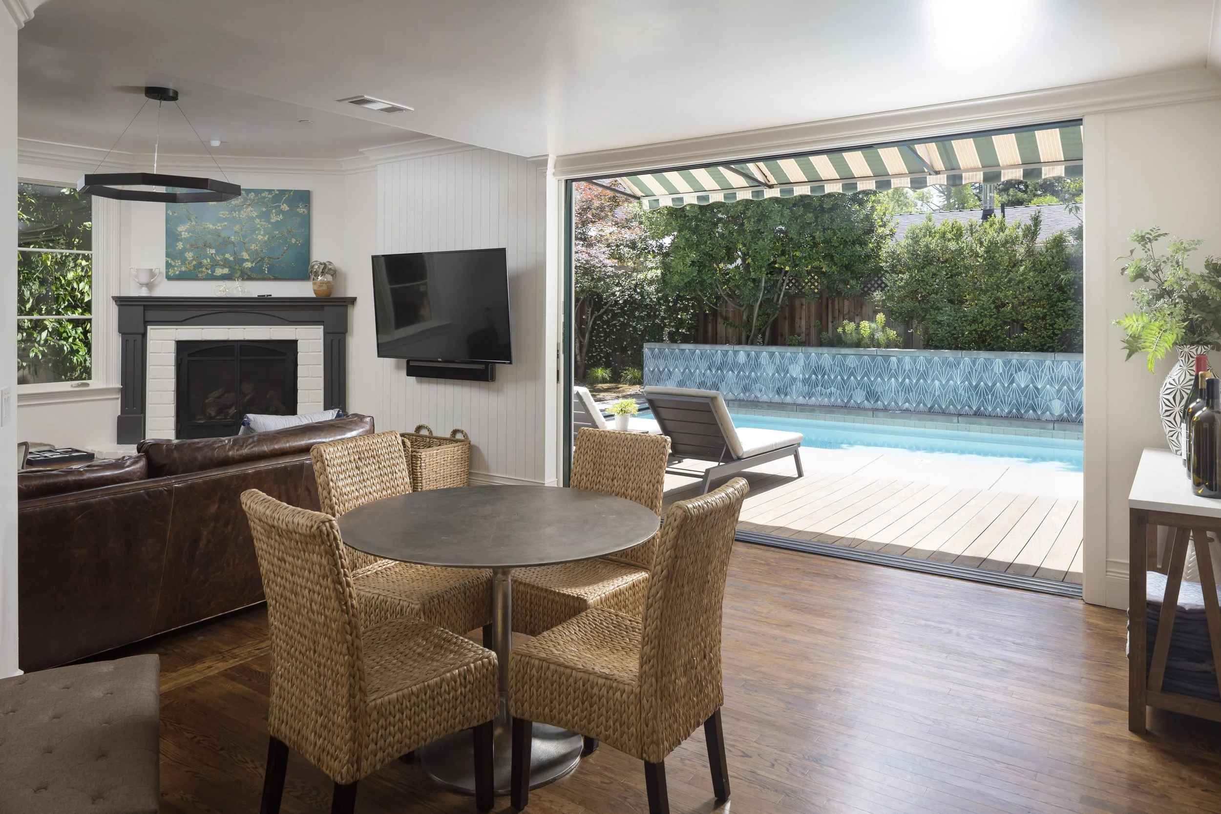 Living room with a fireplace, flat-screen TV, and a dining area with wicker chairs, overlooking a backyard pool with a patterned fence and green trees.