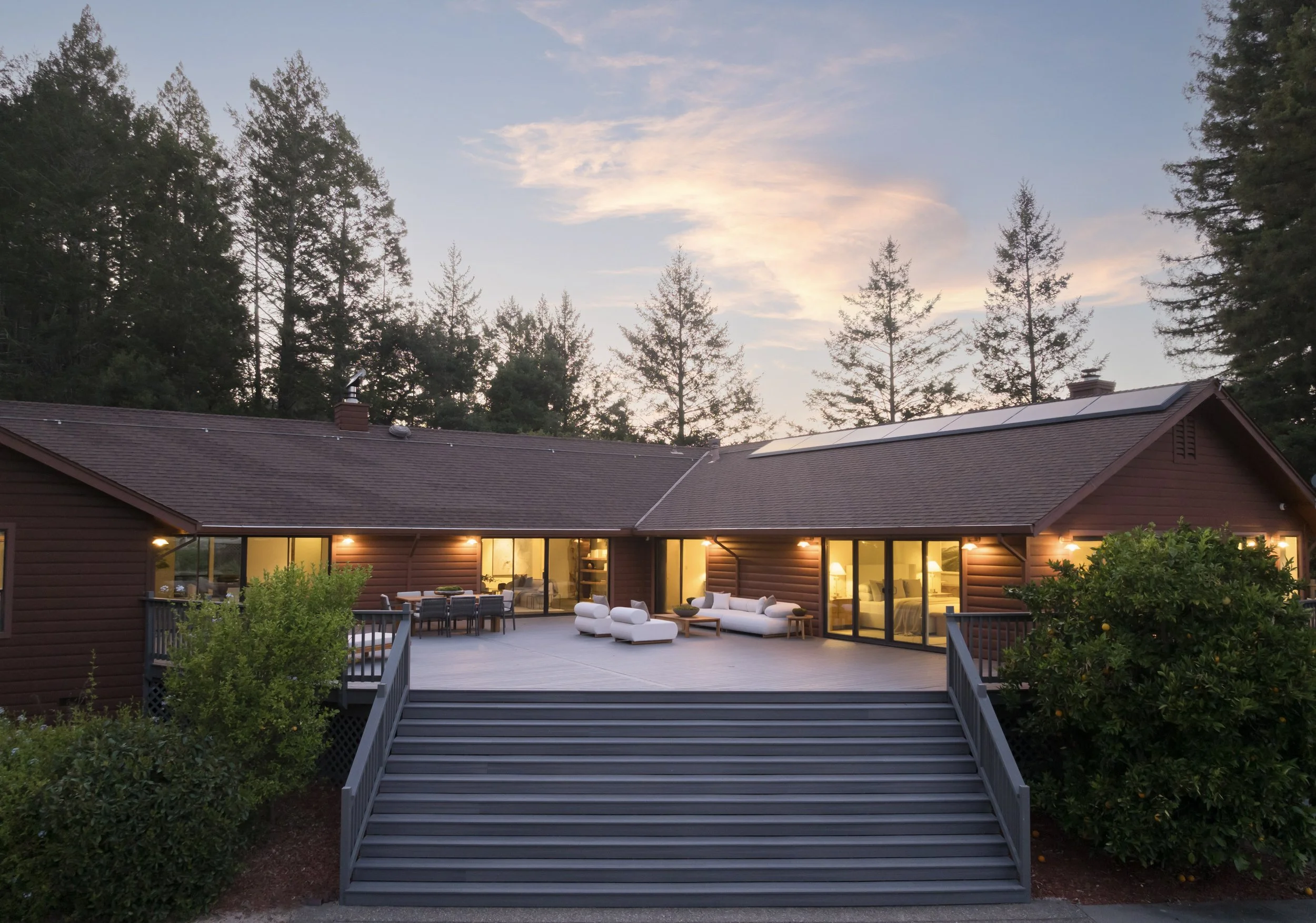 A spacious backyard patio with outdoor furniture, including white cushioned chairs and a dark dining table, leading up to a house with large glass doors. The house has solar panels on the roof and warm interior lighting, surrounded by lush green bush