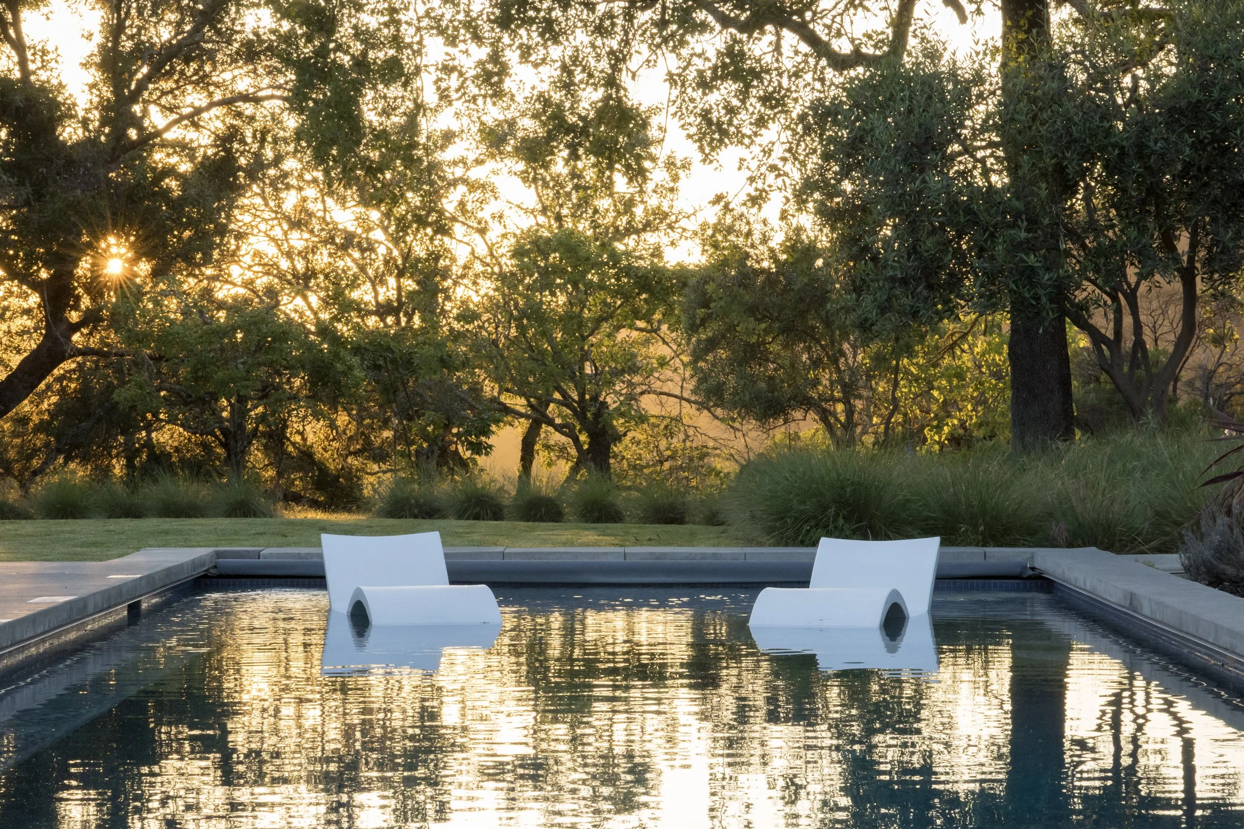 Swimming pool with relaxing white lounge chairs, surrounded by grass and trees, during sunset with sunlight filtering through leaves.