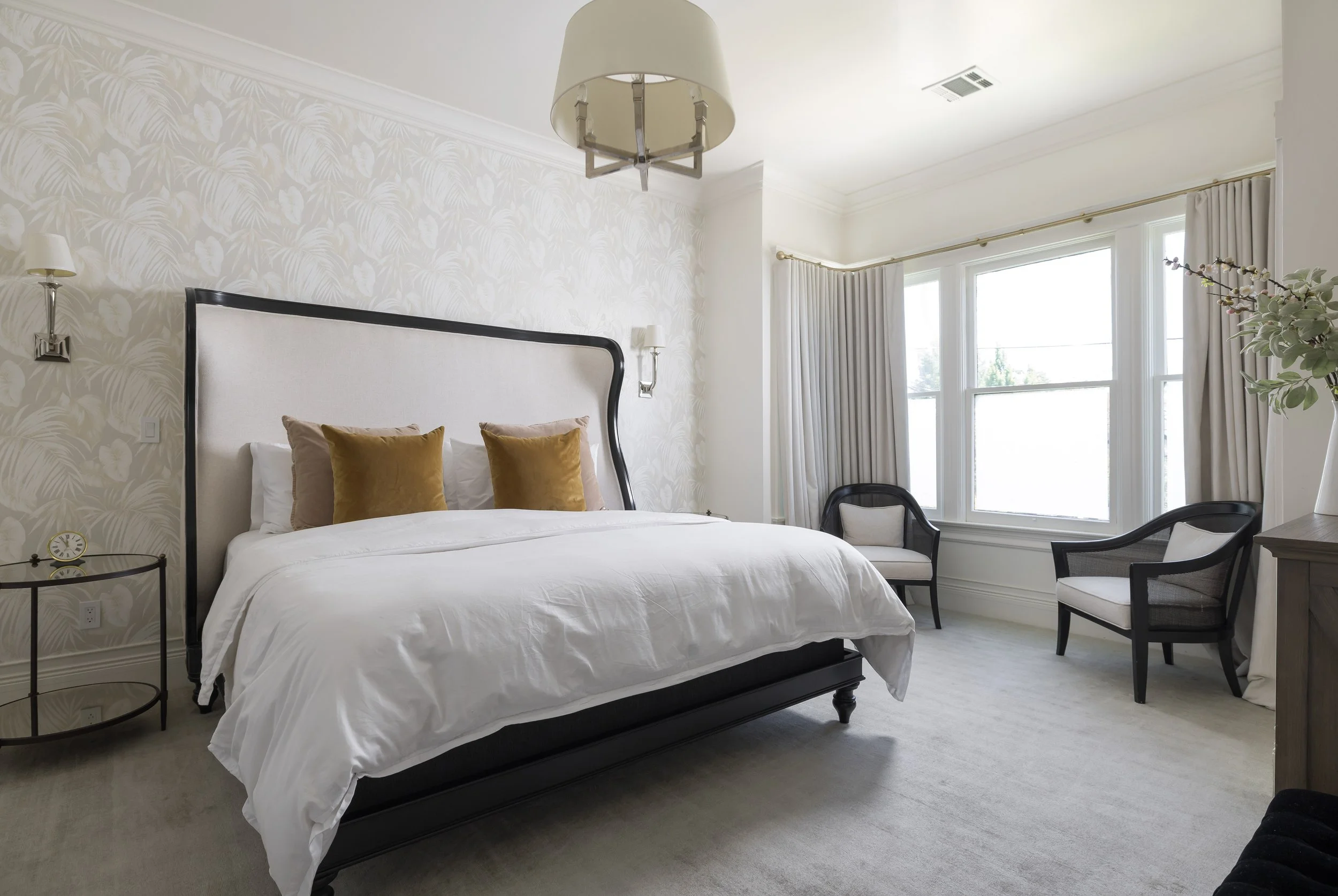 A bedroom with a white bed, brown pillows, beige headboard, side table with a clock, chairs by the window, and a floral wallpaper on the wall. Large windows with curtains and a ceiling lamp overhead.