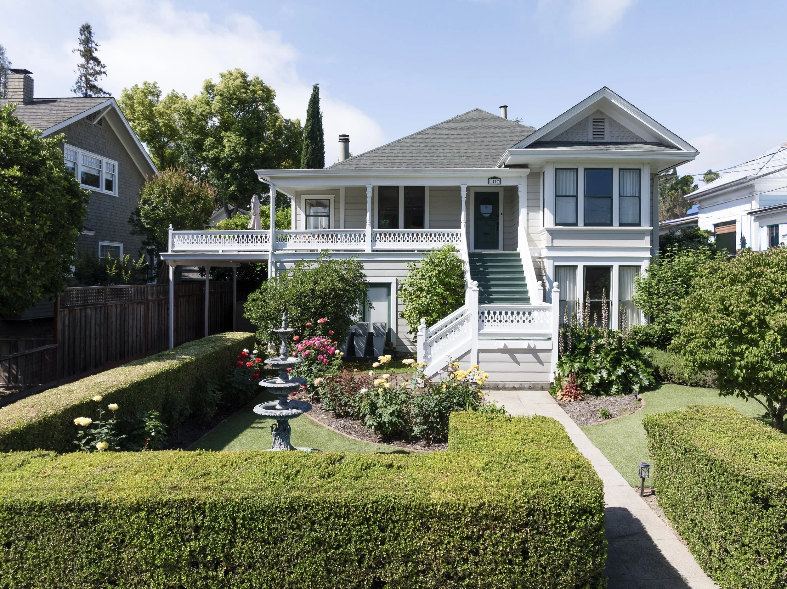 A white house with a front porch, surrounded by a garden with flowers and trimmed bushes, under a partly cloudy sky.