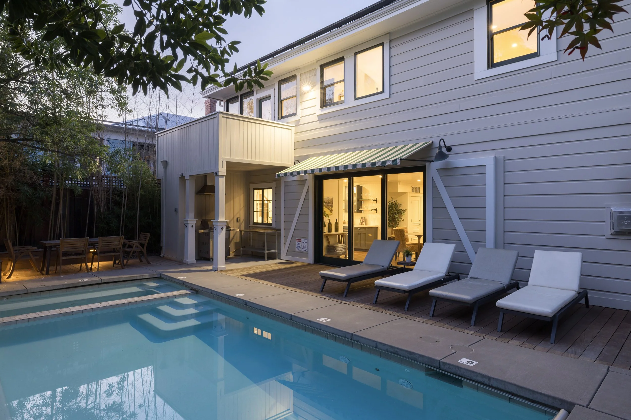 Backyard with swimming pool, lounge chairs, outdoor dining table, and a house with illuminated windows and a striped awning in the evening.