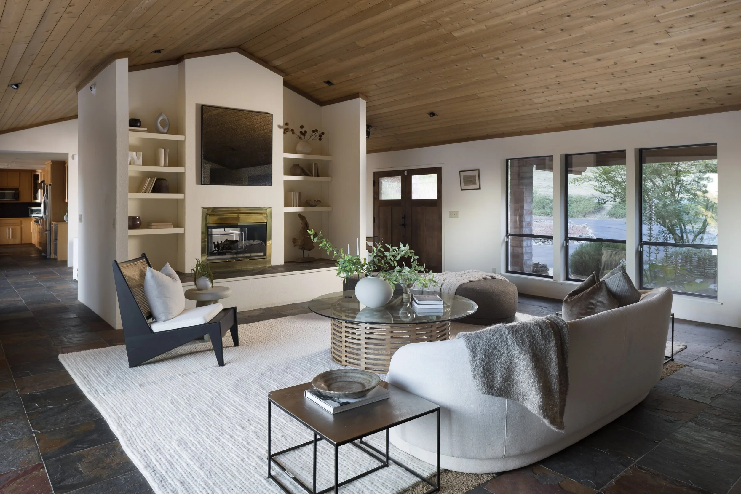 Open living room with a vaulted wooden ceiling, large windows, a fireplace, and modern furniture including a beige sofa, black armchair, and round glass coffee table with decorative vases and books.