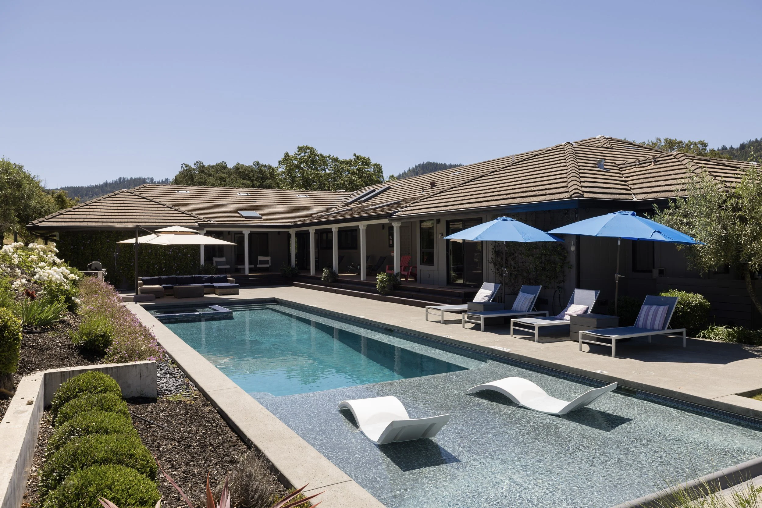 A backyard with a swimming pool, lounge chairs, umbrellas, and a house with a tiled roof and large windows, surrounded by plants and trees, under a clear blue sky.