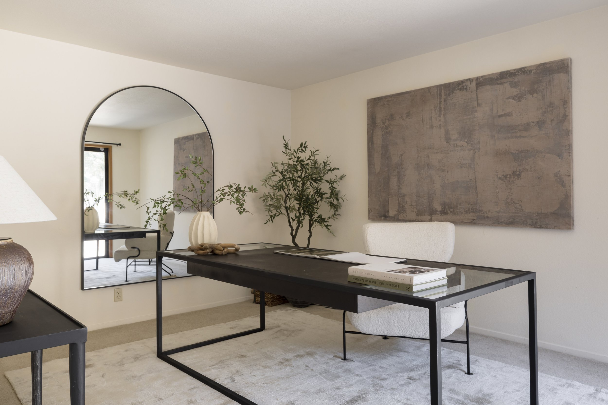 A modern interior with a large mirror, a black desk with books, a white chair, a plant, and abstract wall art.