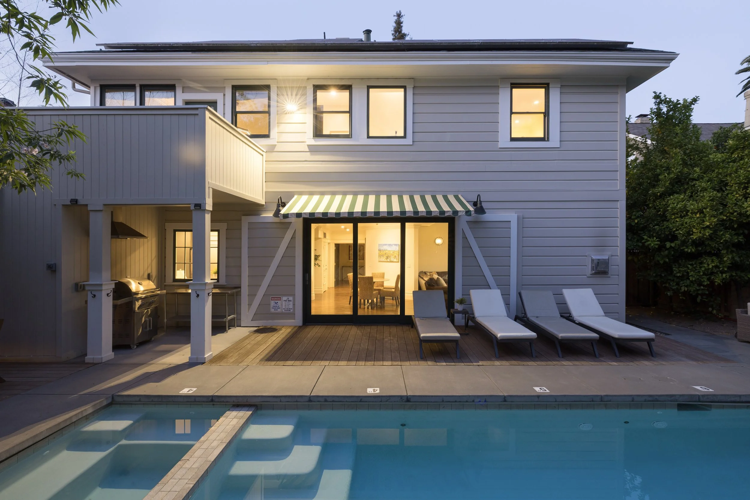 A two-story house with beige siding and large windows, lit from inside, with an outdoor heated pool and lounge chairs, surrounded by trees.