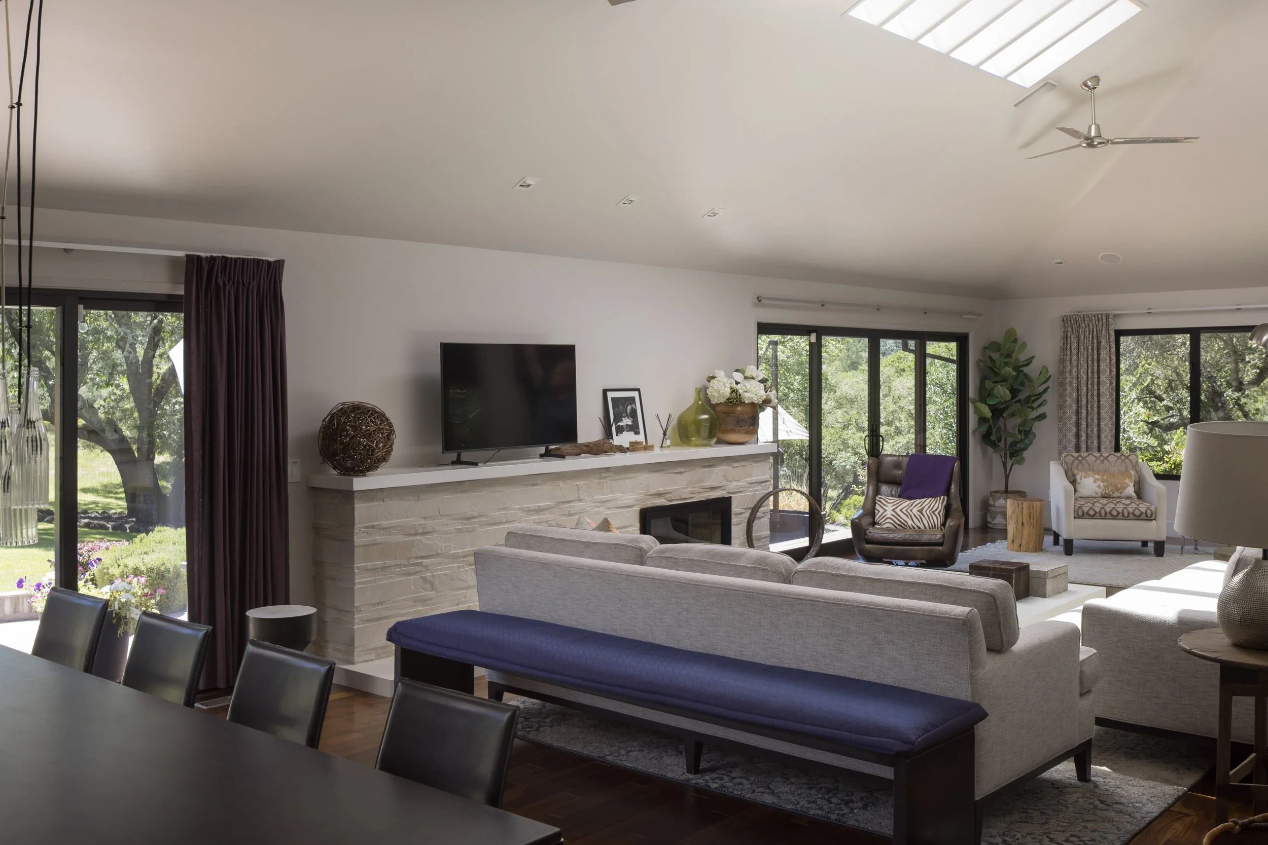 A spacious, modern living room with large windows, a television, decorative vases and framed pictures on a white fireplace mantel, and various chairs and sofas with cushions, all illuminated by natural light from a skylight and glass doors.