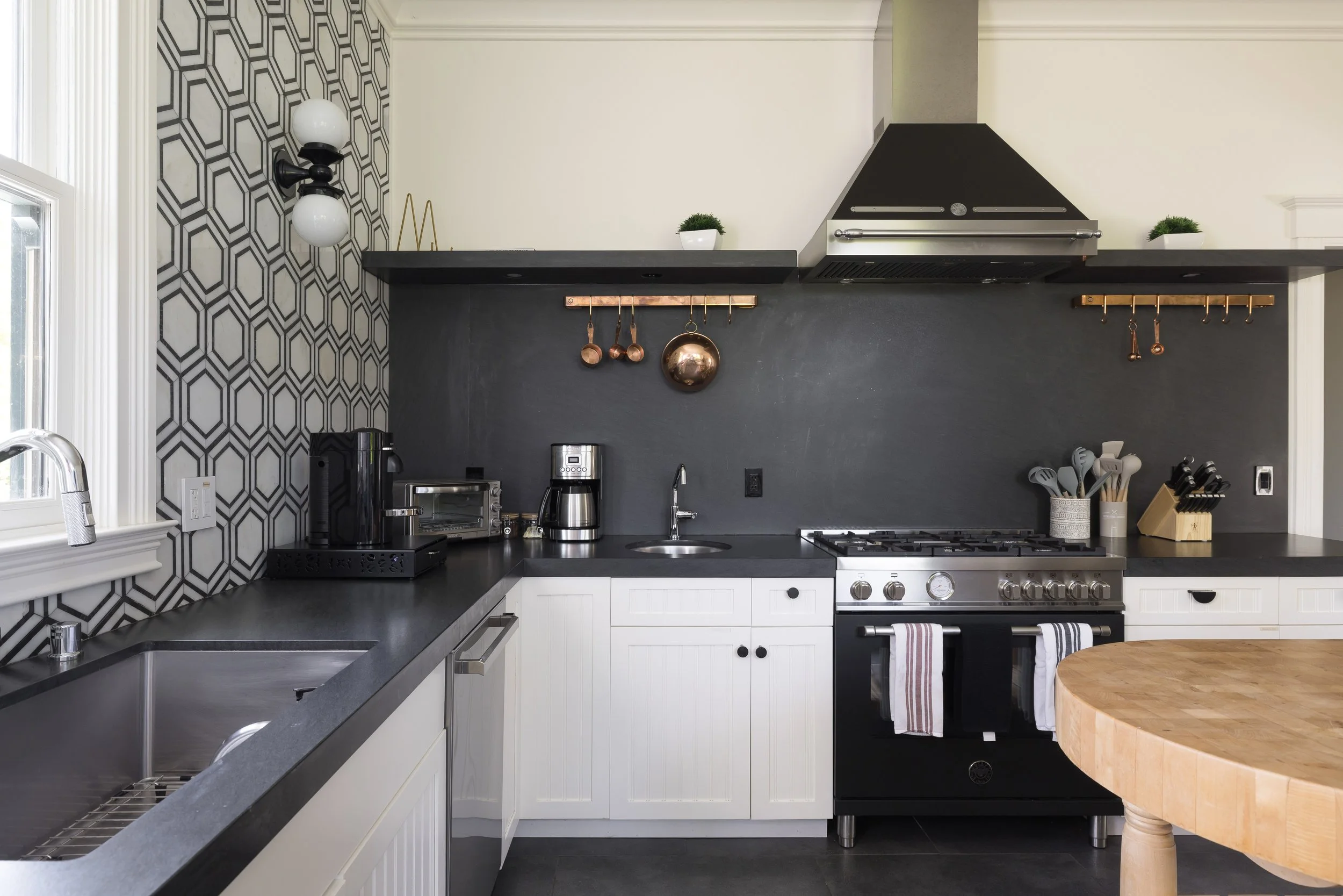 A modern kitchen with black and white cabinets, a black countertop, and a black and white geometric patterned wallpaper. The kitchen has a white sink, coffee maker, toaster oven, and various copper utensils hanging from hooks. There are two small pot