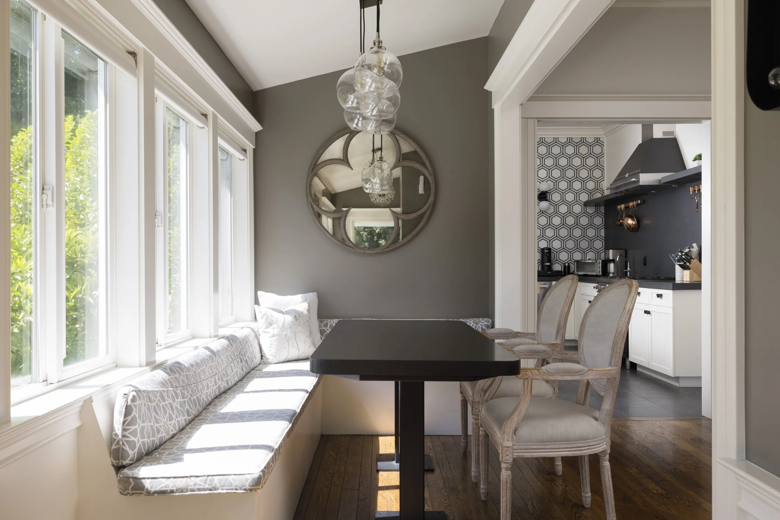 The image shows a cozy dining nook with a long cushioned window seat, a black rectangular dining table, and four beige upholstered chairs. There is a large round mirror with a decorative frame on the grey wall, and three glass pendant lights hanging 