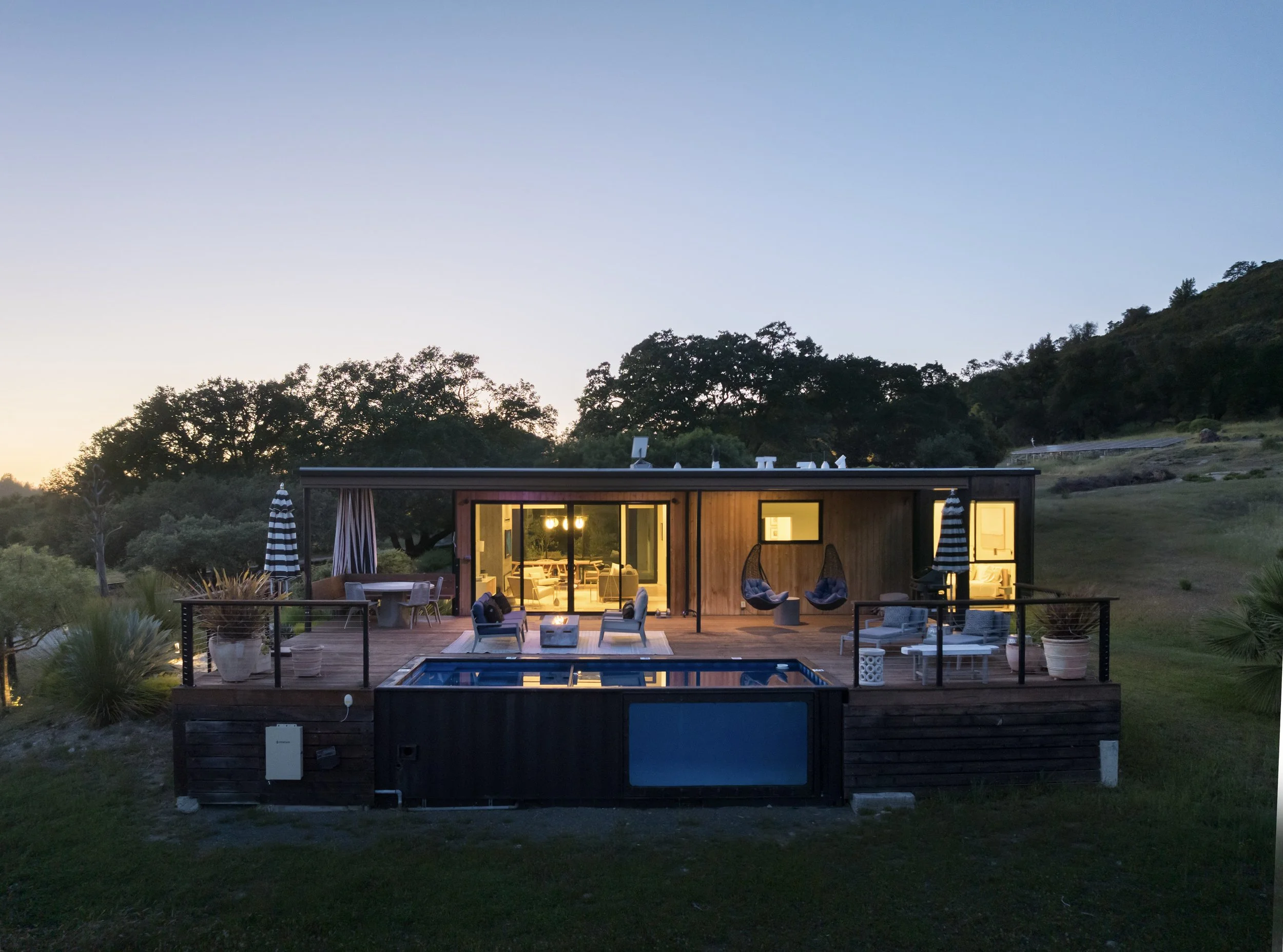 A nighttime scene of a house with large windows, illuminated from inside, surrounded by trees and a hillside, with outdoor furniture, a small swimming pool, and umbrellas on a deck.