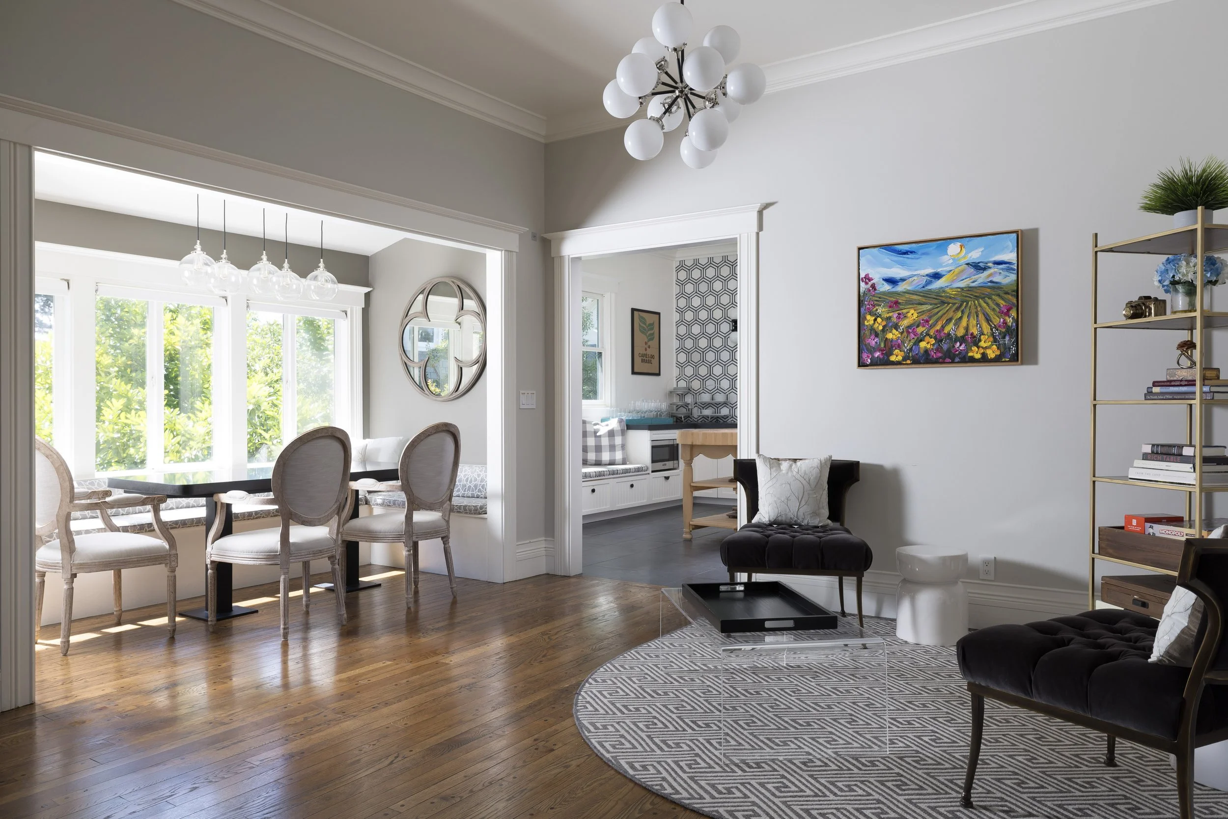 A bright living and dining area with a hardwood floor, gray walls, white crown molding, and large windows. The dining area has a wooden table with six upholstered chairs, and a modern chandelier with globe lights. The living space has a black tufted 