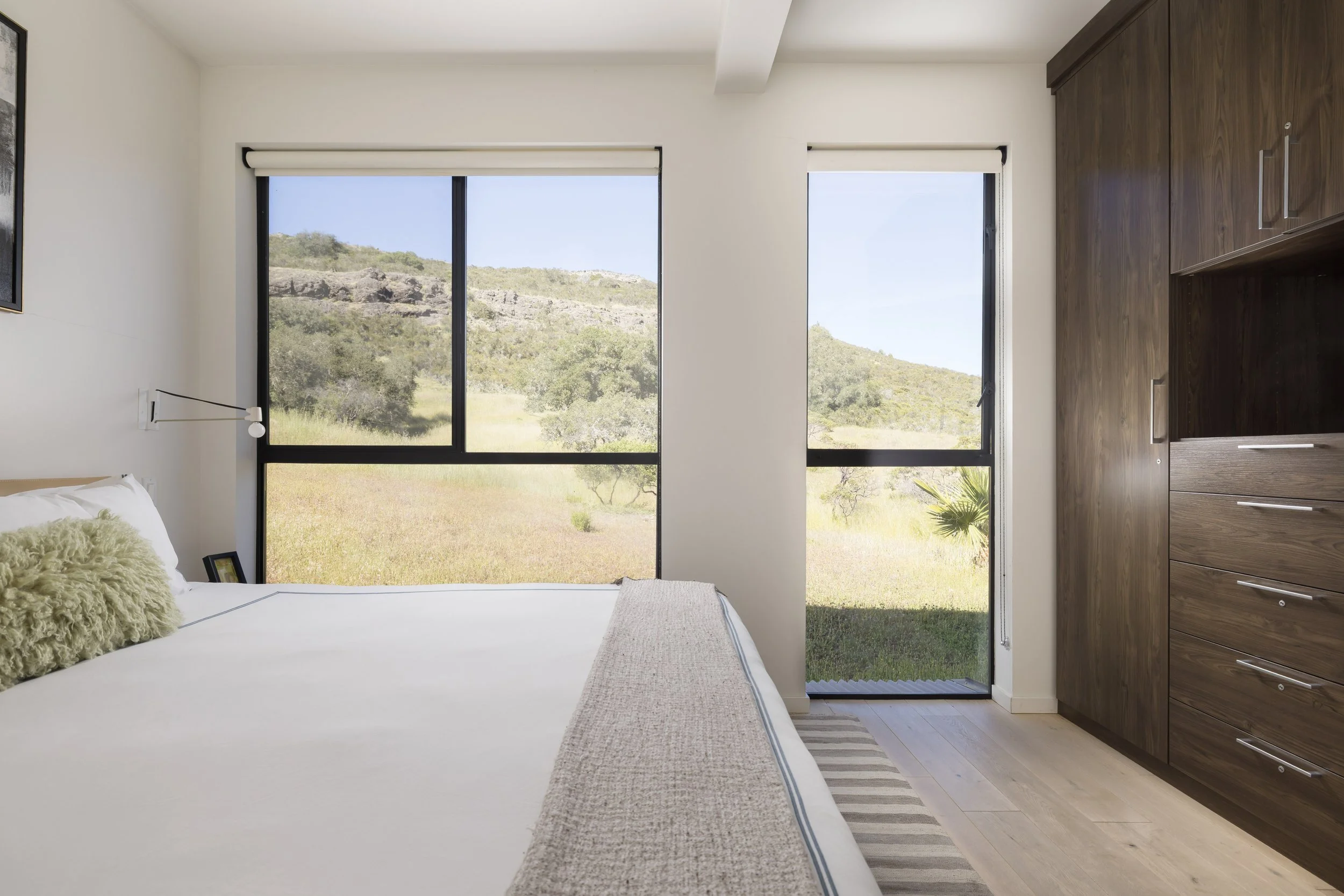 View from inside a bedroom showing large windows with a landscape of green hills and trees outside, a wooden dresser on the right, and part of a bed with pillows on the left.