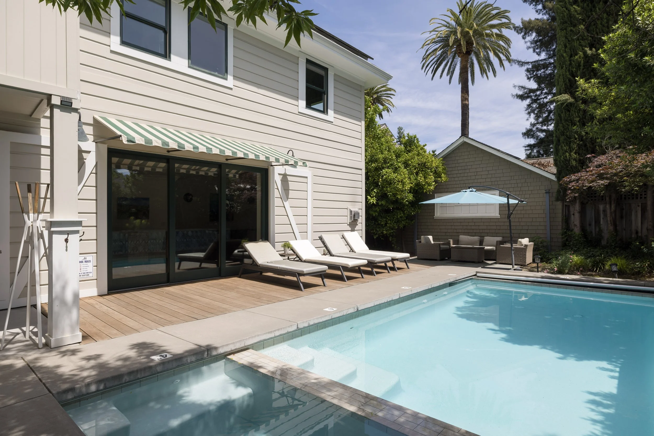 A backyard swimming pool with lounge chairs, a patio, an umbrella, and a house with large windows and a striped awning, surrounded by trees including a tall palm tree.