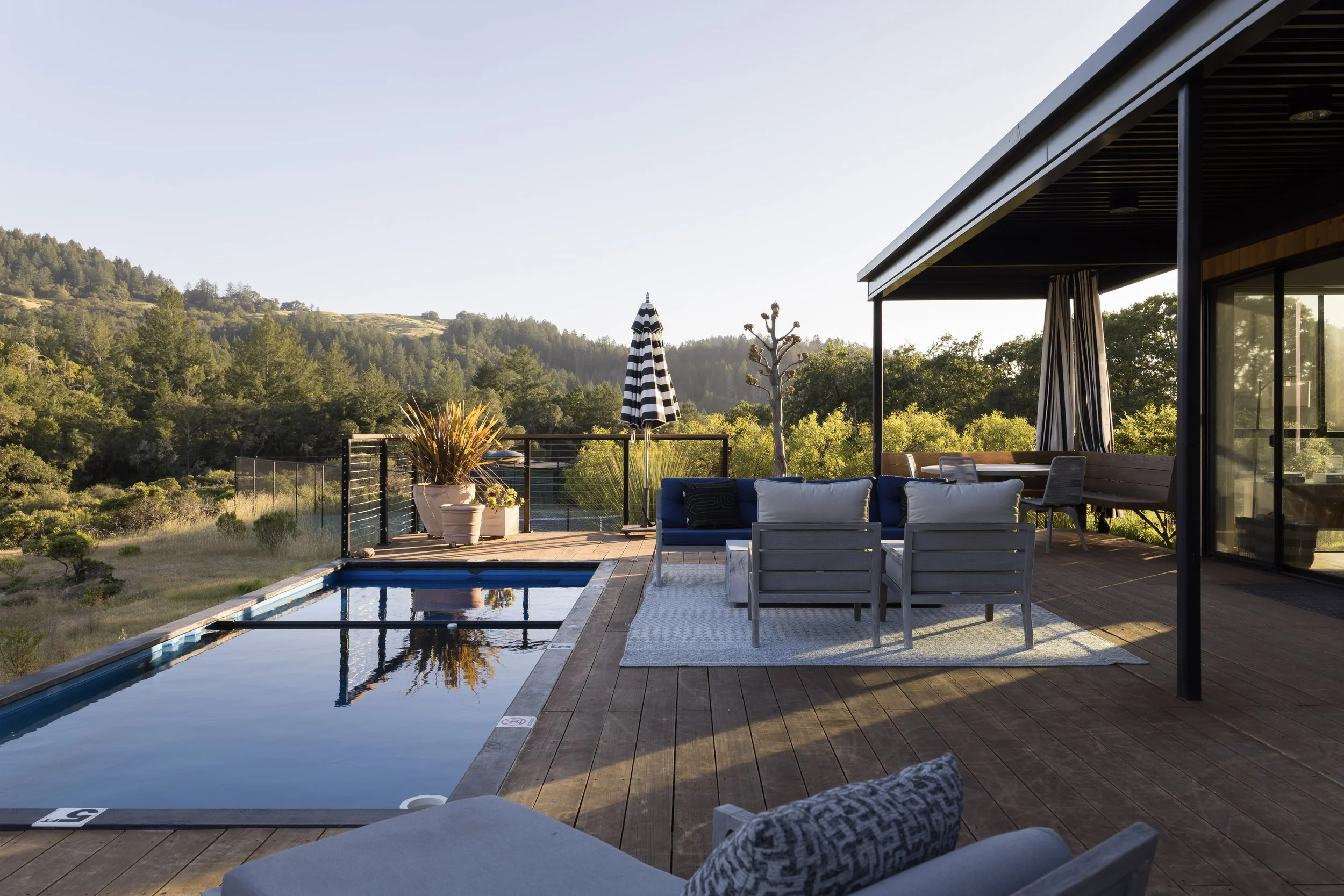A backyard patio with a swimming pool, outdoor furniture, a striped umbrella, and trees overlooking a hillside in the distance.