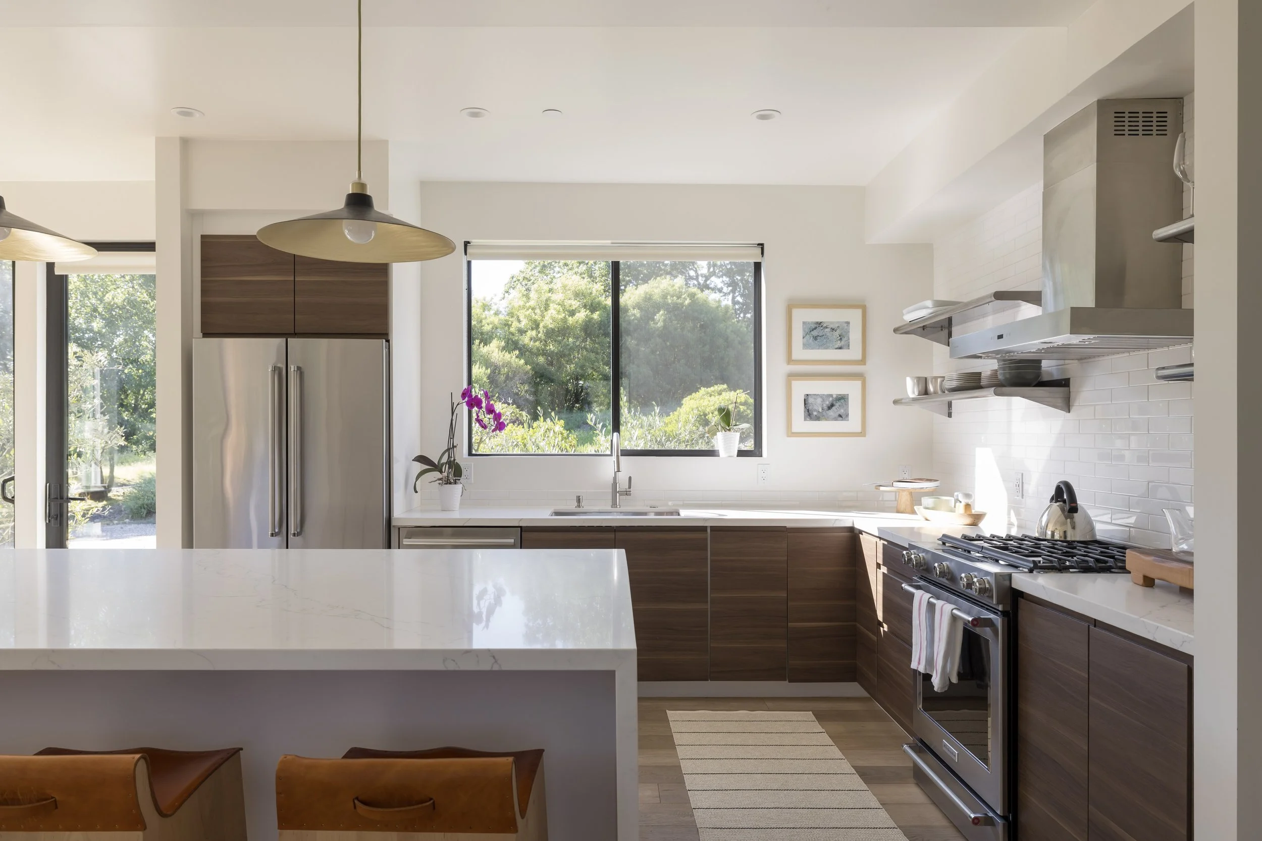 Modern kitchen with stainless steel appliances, dark wood cabinets, white countertops, large window with view of greenery, purple orchids on the counter, and natural light.