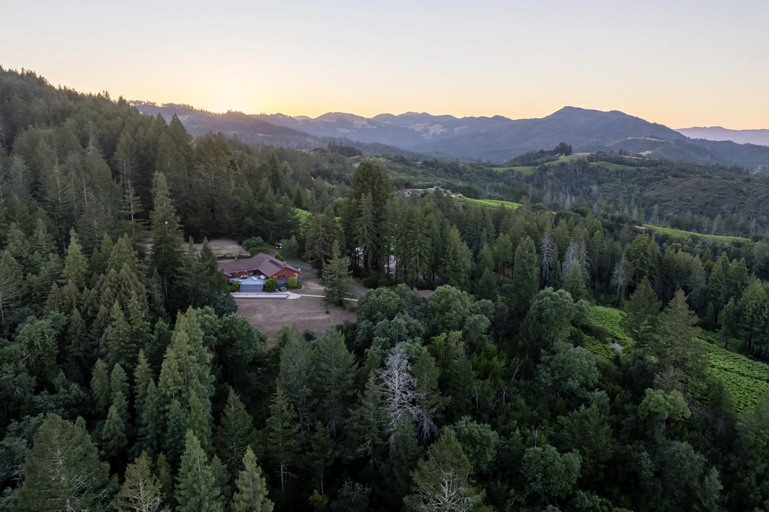 A scenic view of a forested mountainous landscape during sunset with a house and fields in the foreground.