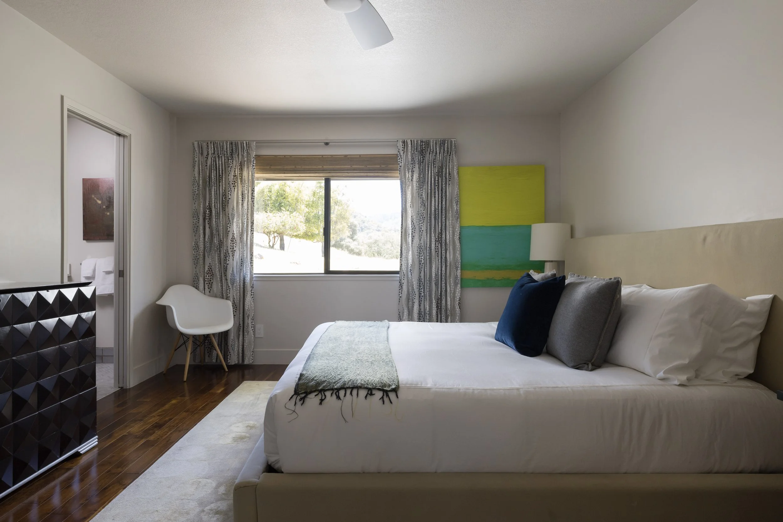 A bedroom with a large window showing trees outside, decorated with patterned curtains. The bed has a beige headboard, white bedding, and several pillows, including a dark blue one. There is a colorful abstract painting on the wall and a nightstand w