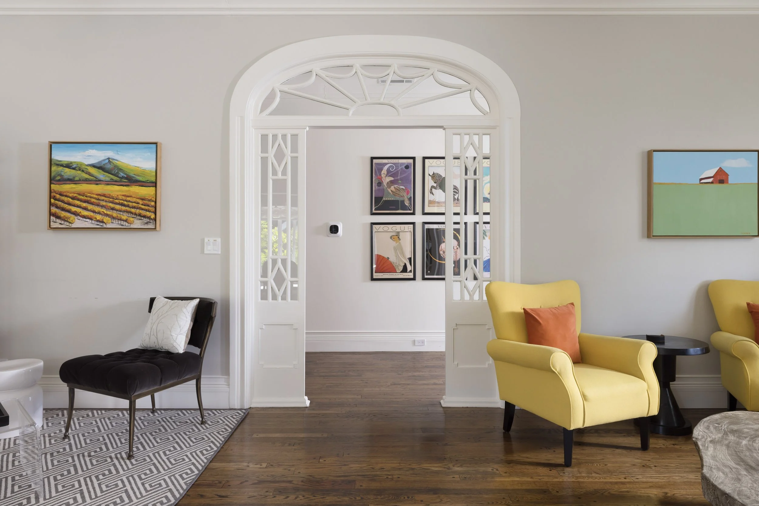 A bright living room with gray walls, featuring colorful artwork and furniture, including a black chair with a white pillow, yellow armchairs with orange pillows, and a patterned rug on wooden flooring.