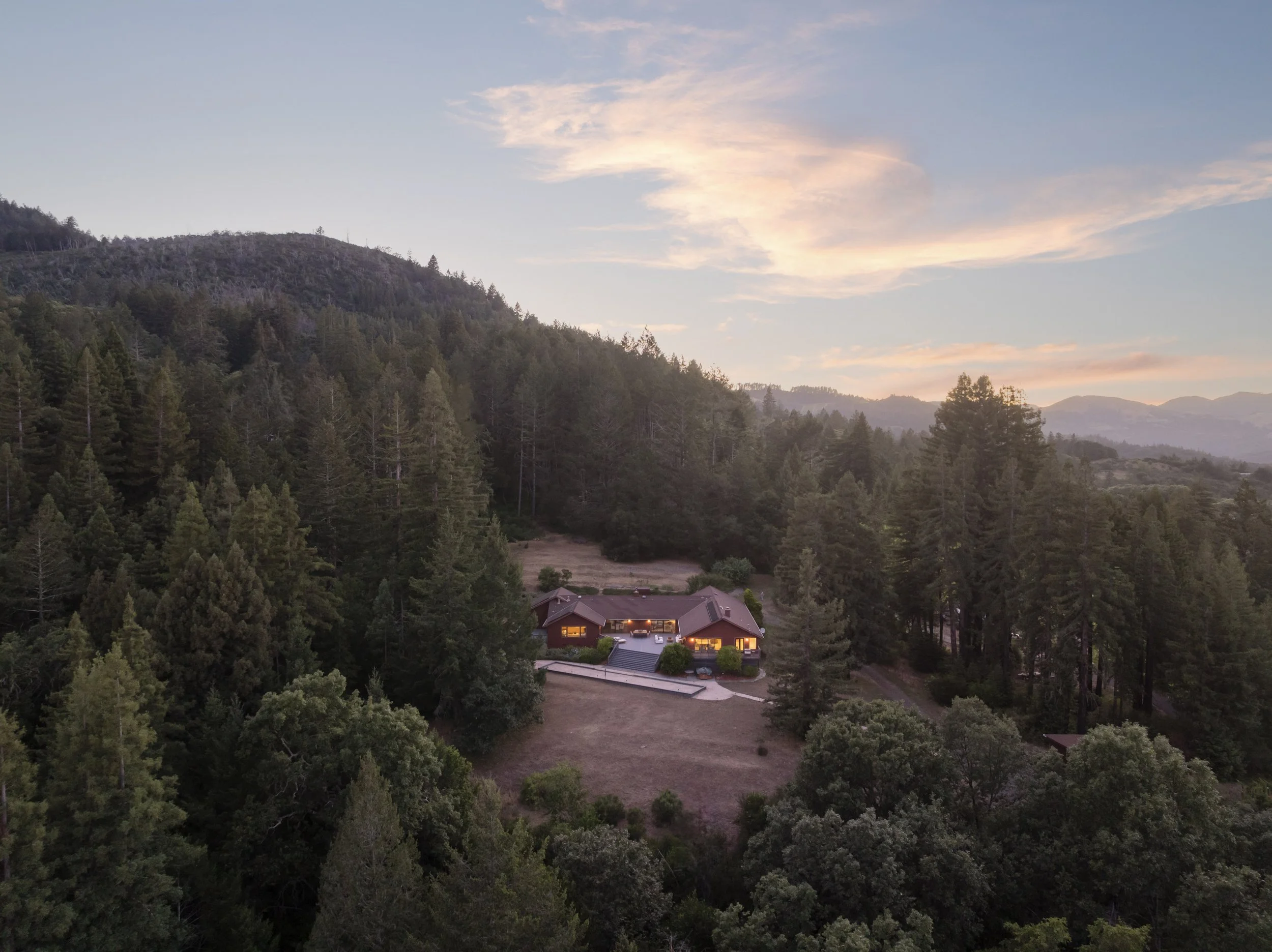 A house nestled in a forested hillside at dusk, with the sky displaying soft pink and orange clouds.
