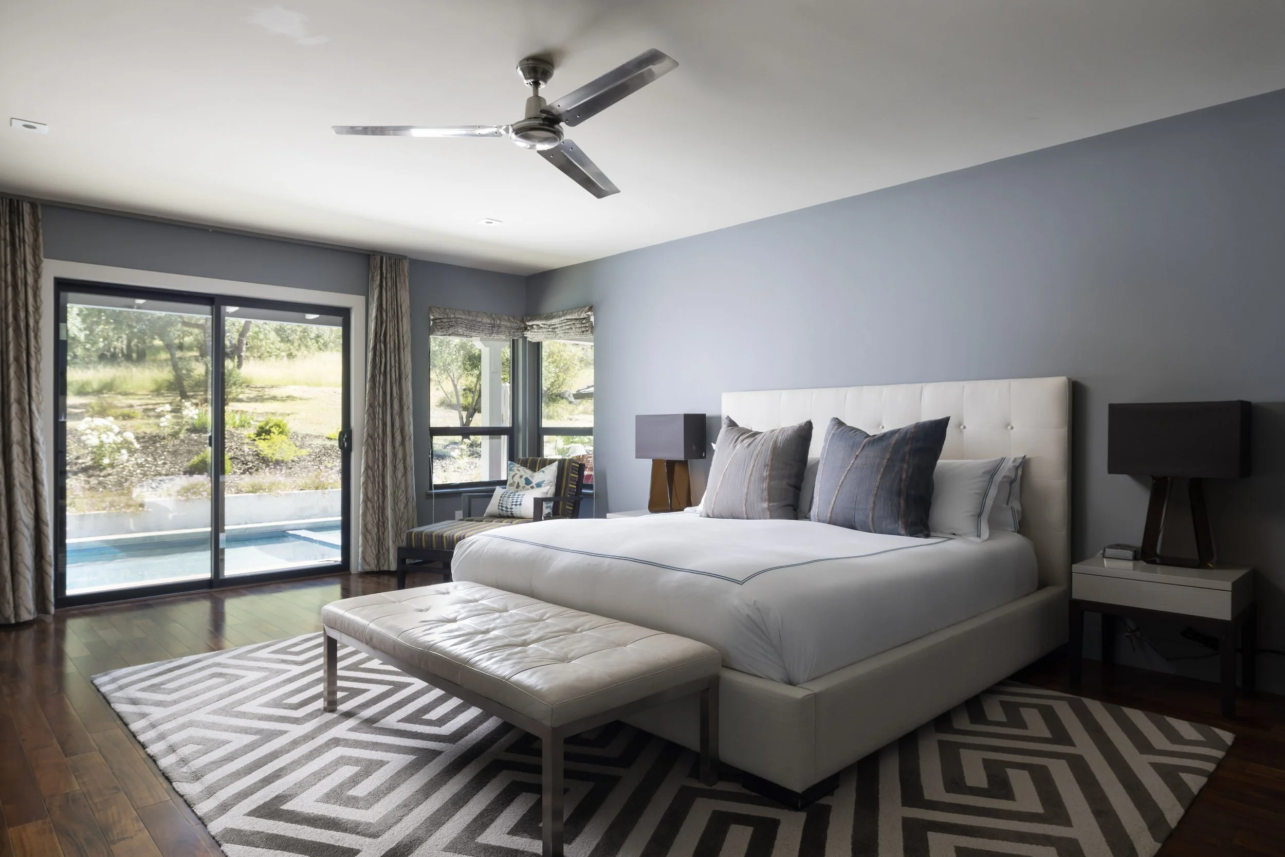 Contemporary bedroom with a sliding glass door leading to an outdoor patio, a white bed with gray pillows, a bench at the foot of the bed, dark nightstands on either side, a ceiling fan, and windows with patterned valances, all illuminated by natural