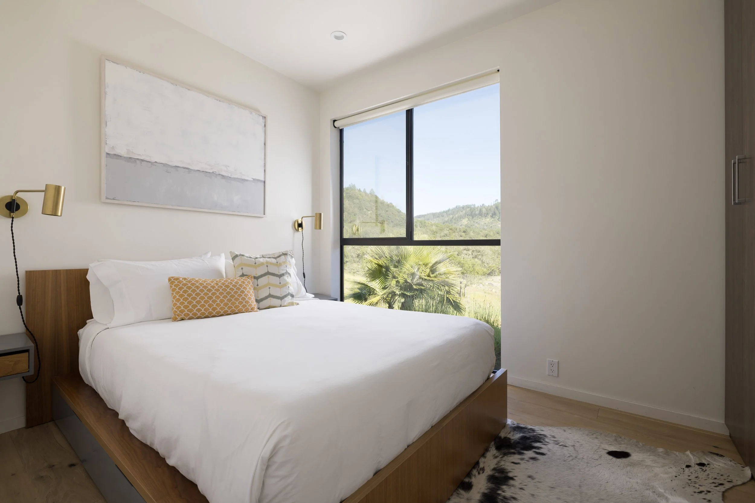 A modern bedroom with a large bed, decorative pillows, wall-mounted reading lamps, a large window showing a view of green hills and trees, an abstract wall art piece, hardwood flooring, and a cowhide rug.