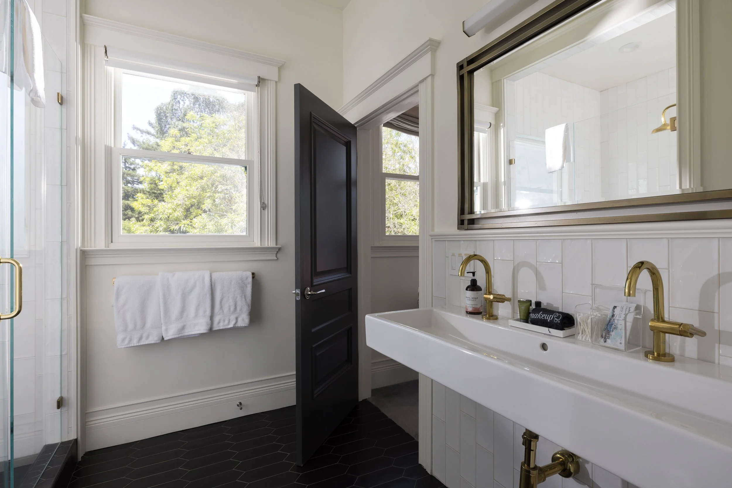 Bright bathroom with white walls and trim, a large mirror, black door, window with trees outside, white sink with gold fixtures, and various toiletries.