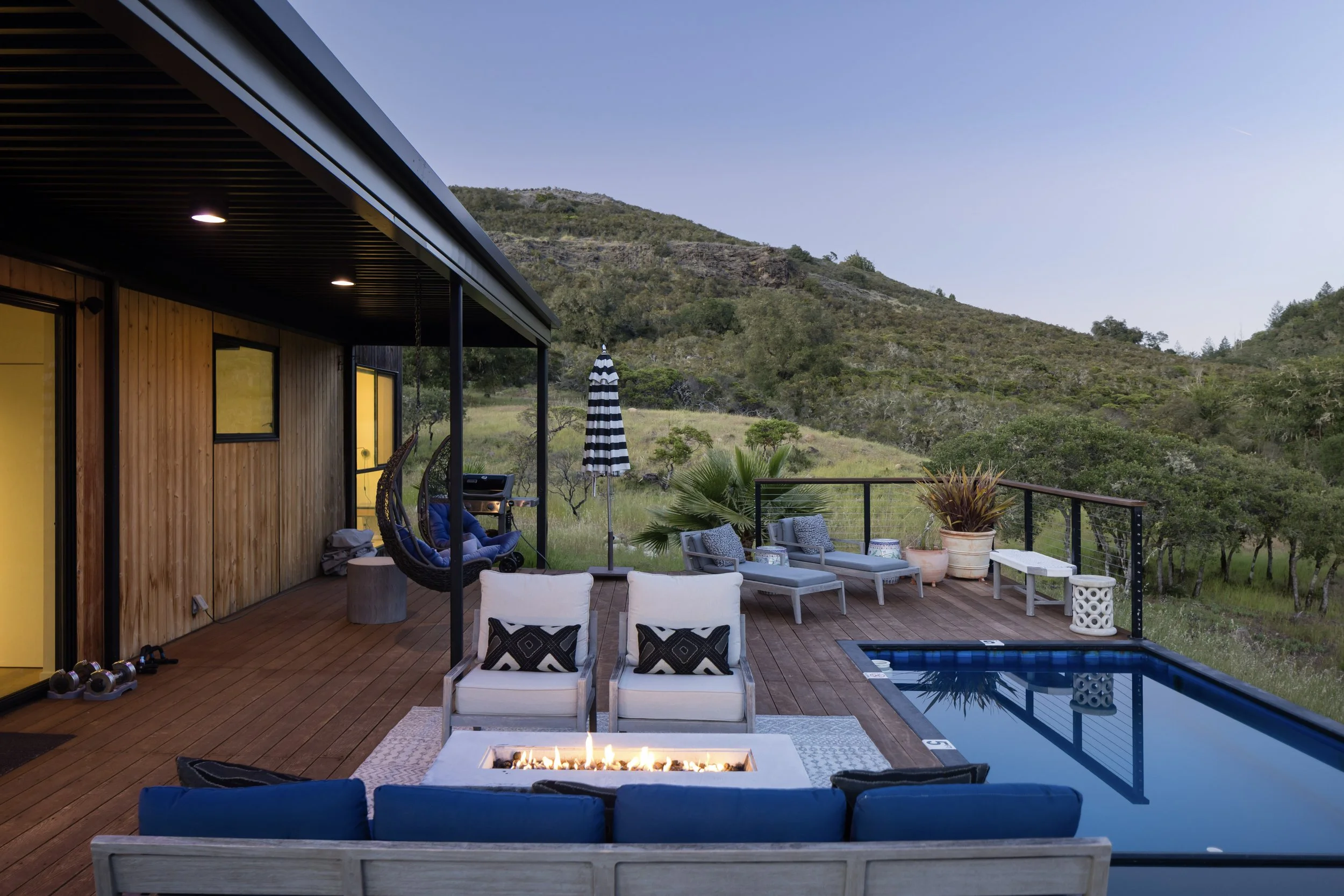 Outdoor deck with seating, a small pool, a fire pit, potted plants, and mountain scenery in the background.