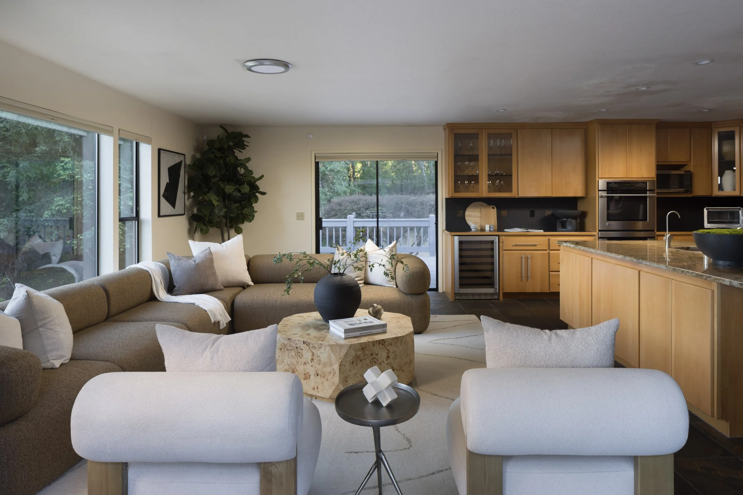 Living room with beige sofa, white armchairs, black side table with geometric white sculpture, marble coffee table, large windows showing outdoor greenery, kitchen with wooden cabinets, black backsplash, and stainless steel appliances.