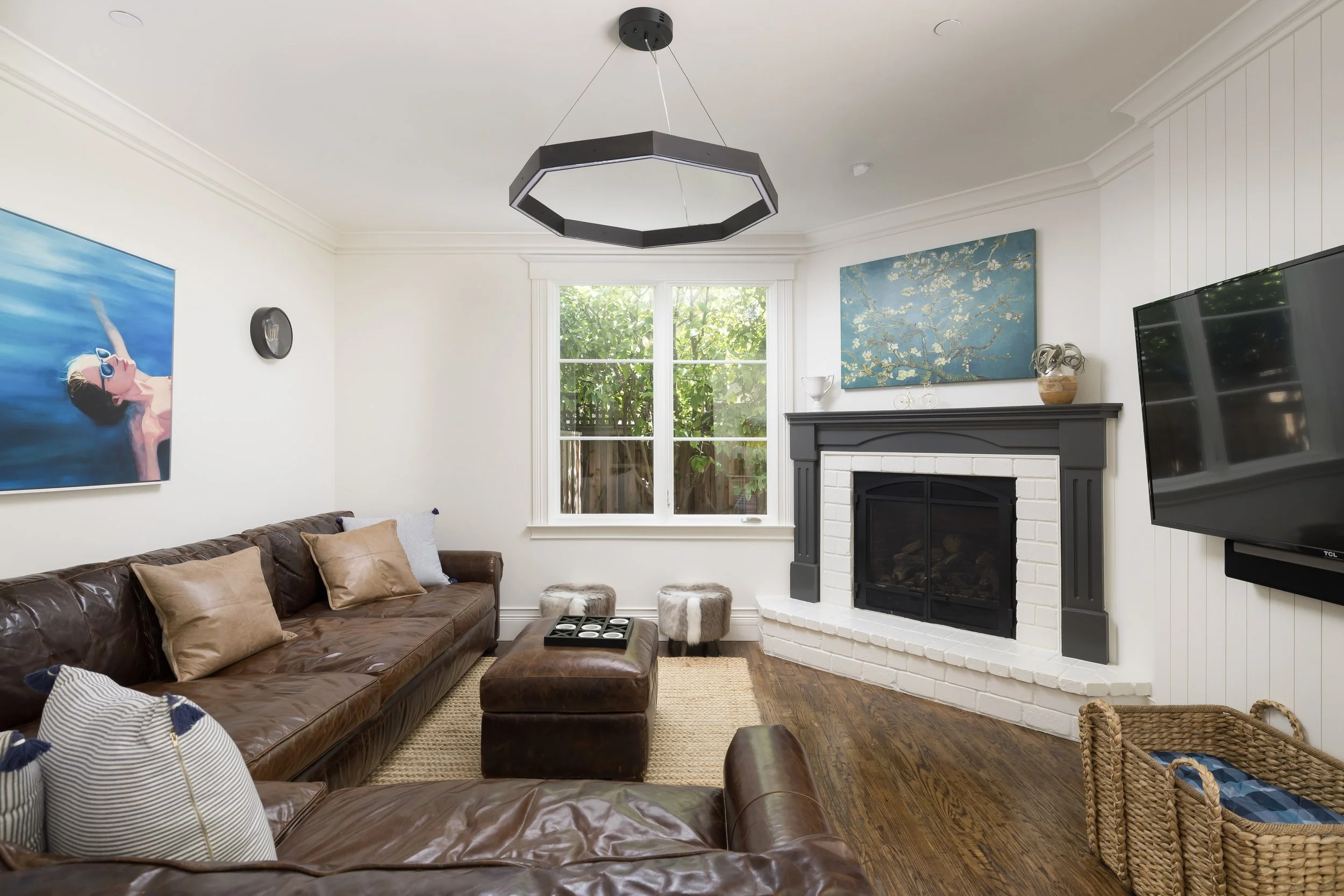 Living room with a brown leather sofa, a black fireplace, two small white stools, a TV mounted on the wall, and artwork on the walls, with a window showing green trees outside.