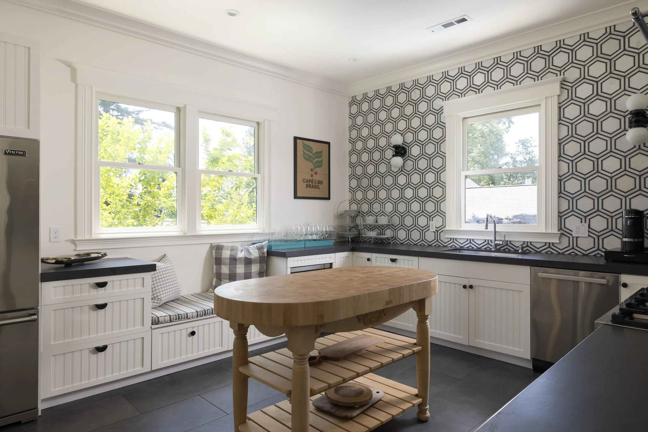 A modern kitchen with white cabinets, black countertops, and a geometric patterned backsplash. There are two large windows letting in natural light, with green trees visible outside. The kitchen features a round wooden table, a small bench with pillo