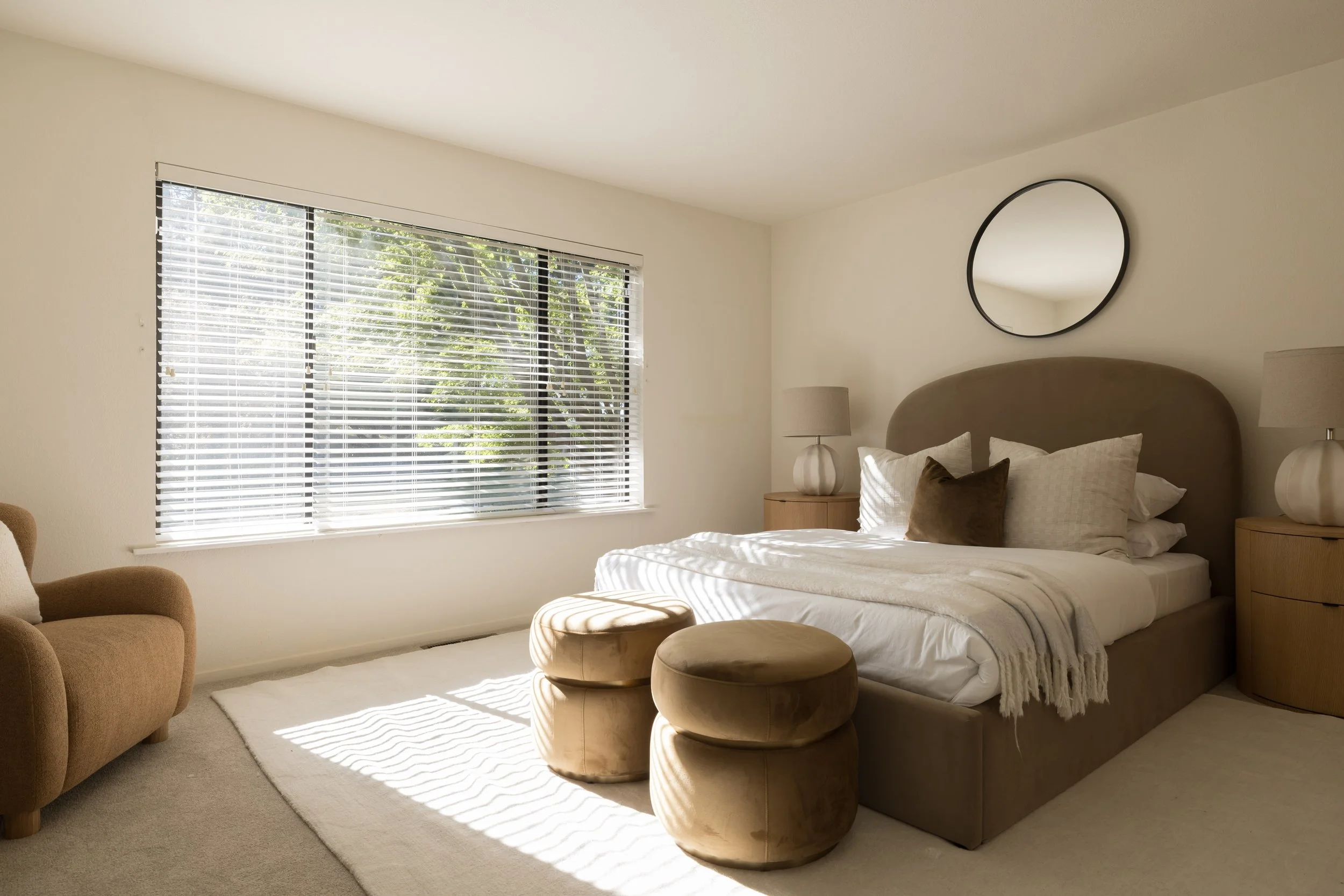 A bedroom with a large window with blinds, a bed with white linens and pillows, a round mirror on the wall, two beige lamps on bedside tables, and tan ottomans at the foot of the bed.