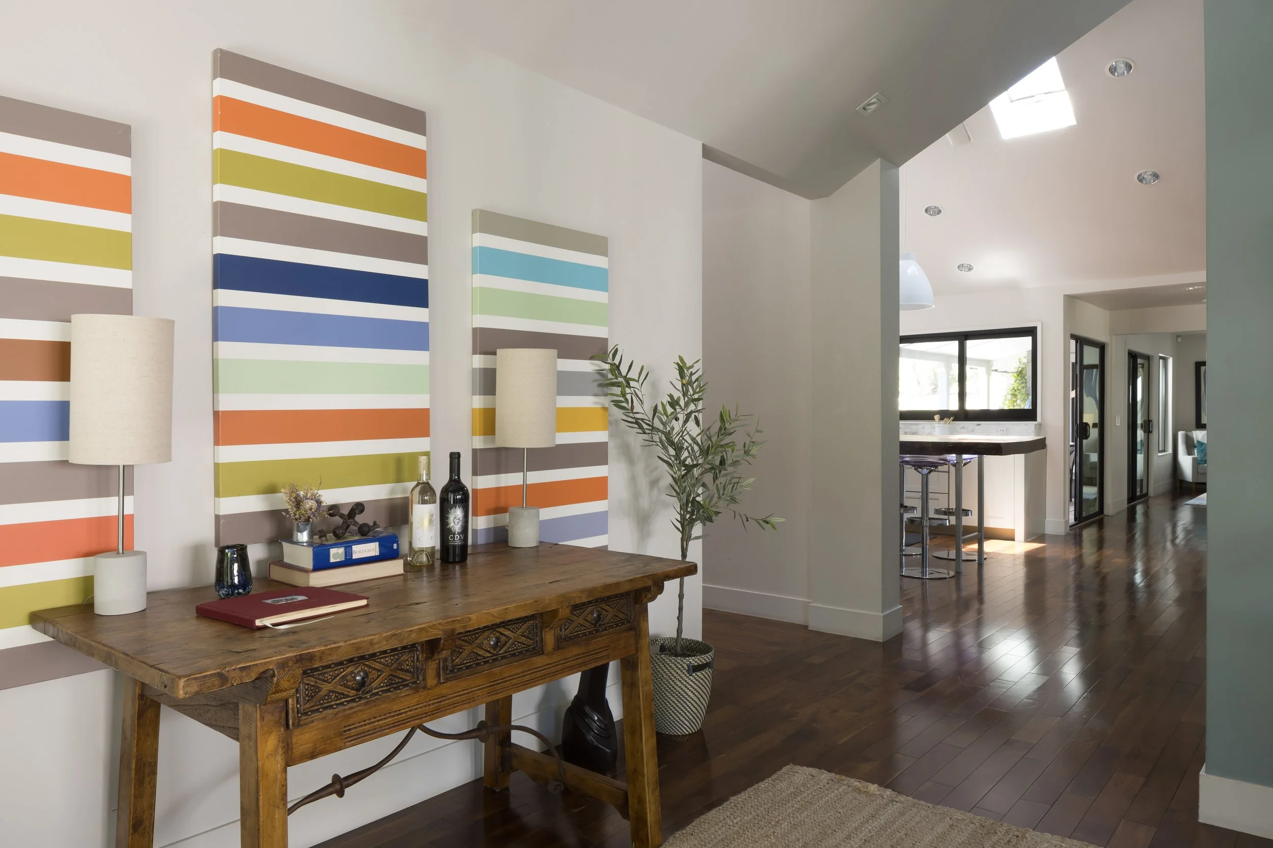 Interior view of a modern living space featuring artwork with colorful horizontal stripes, wooden furniture, decorative items, and a hallway leading to a kitchen with large windows.