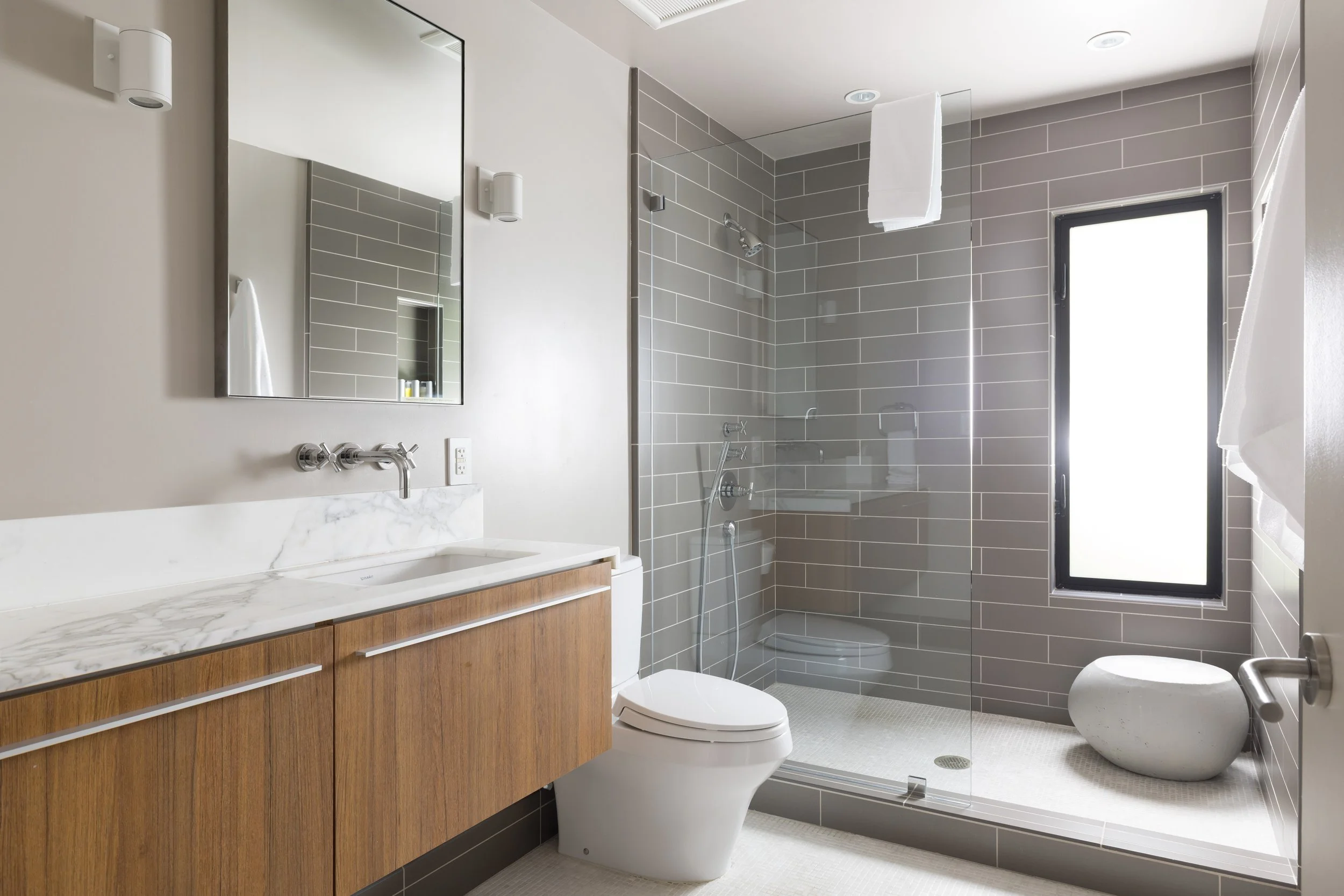 Modern bathroom with a wooden vanity, marble countertop, wall-mounted mirror, toilet, and a glass-enclosed shower with gray tiles and a frosted window.