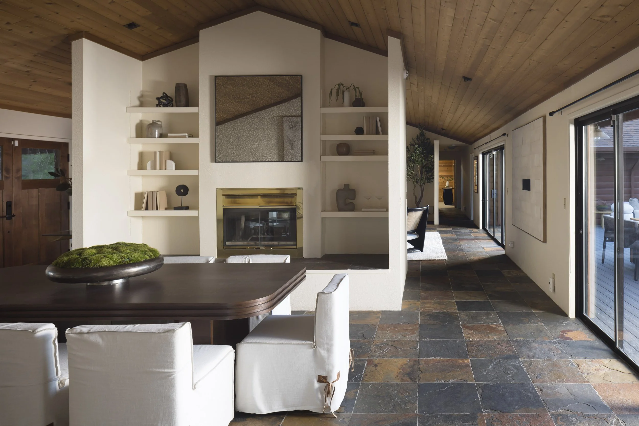 Interior view of a living room with a wooden vaulted ceiling, built-in white shelves around a fireplace, and large sliding glass doors leading to an outdoor patio.
