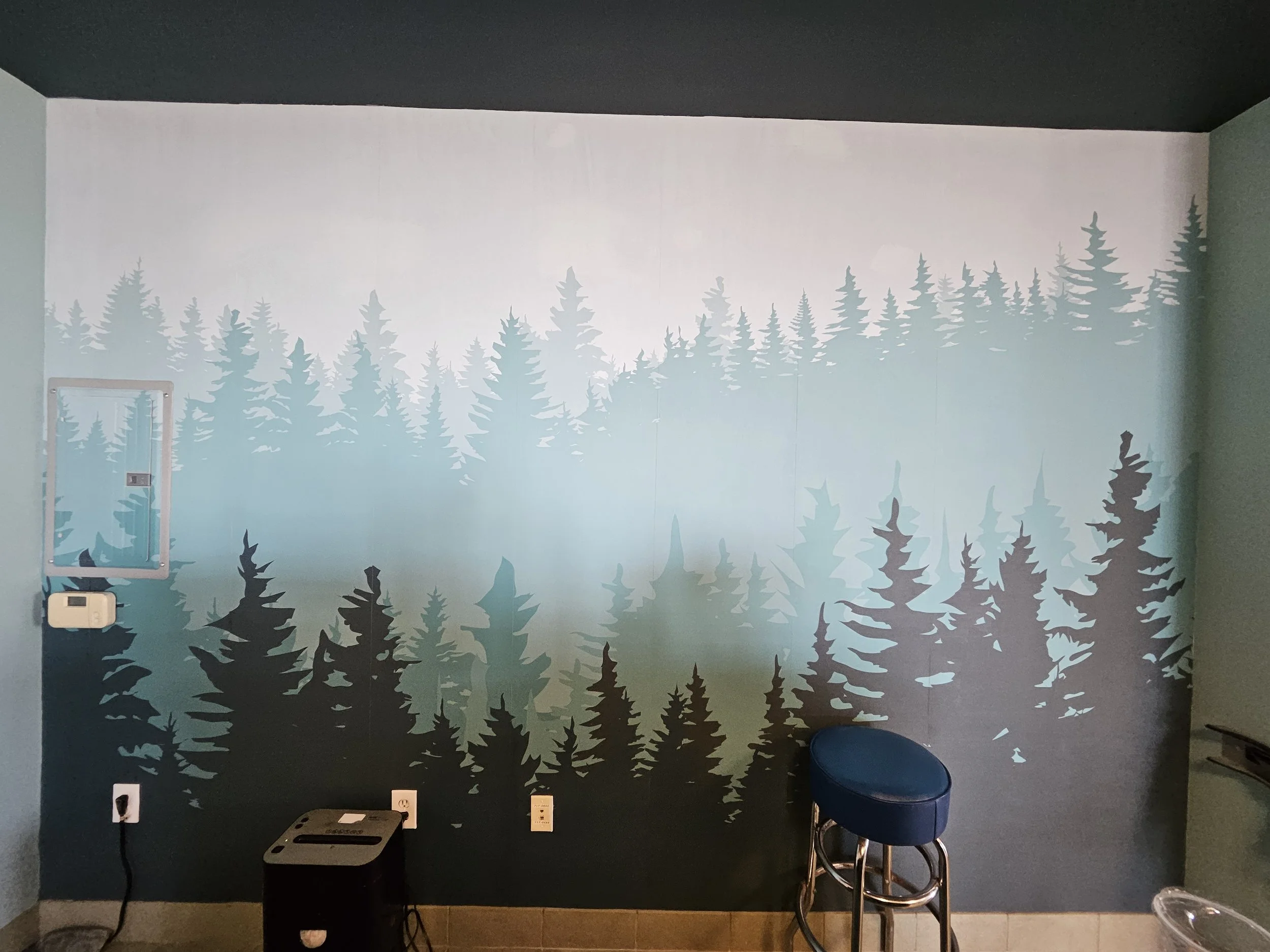 A wall decorated with a mural of silhouettes of pine trees in various shades of blue and green, creating a forest scene. There is a blue stool in front of the mural and a small black table with some electronic devices on it. The room has electrical outlets and a mirror mounted on the wall.