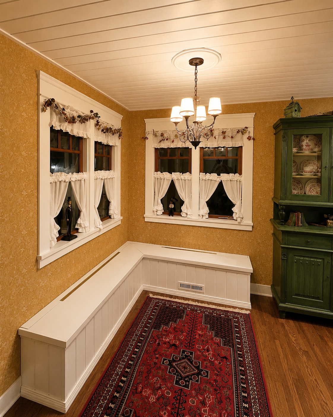 A cozy dining nook corner with three windows dressed in white curtains and decorated with autumn-themed garlands, a white built-in bench, a green china cabinet with dishes, a chandelier hanging from the ceiling, and a red patterned rug on the wooden floor.