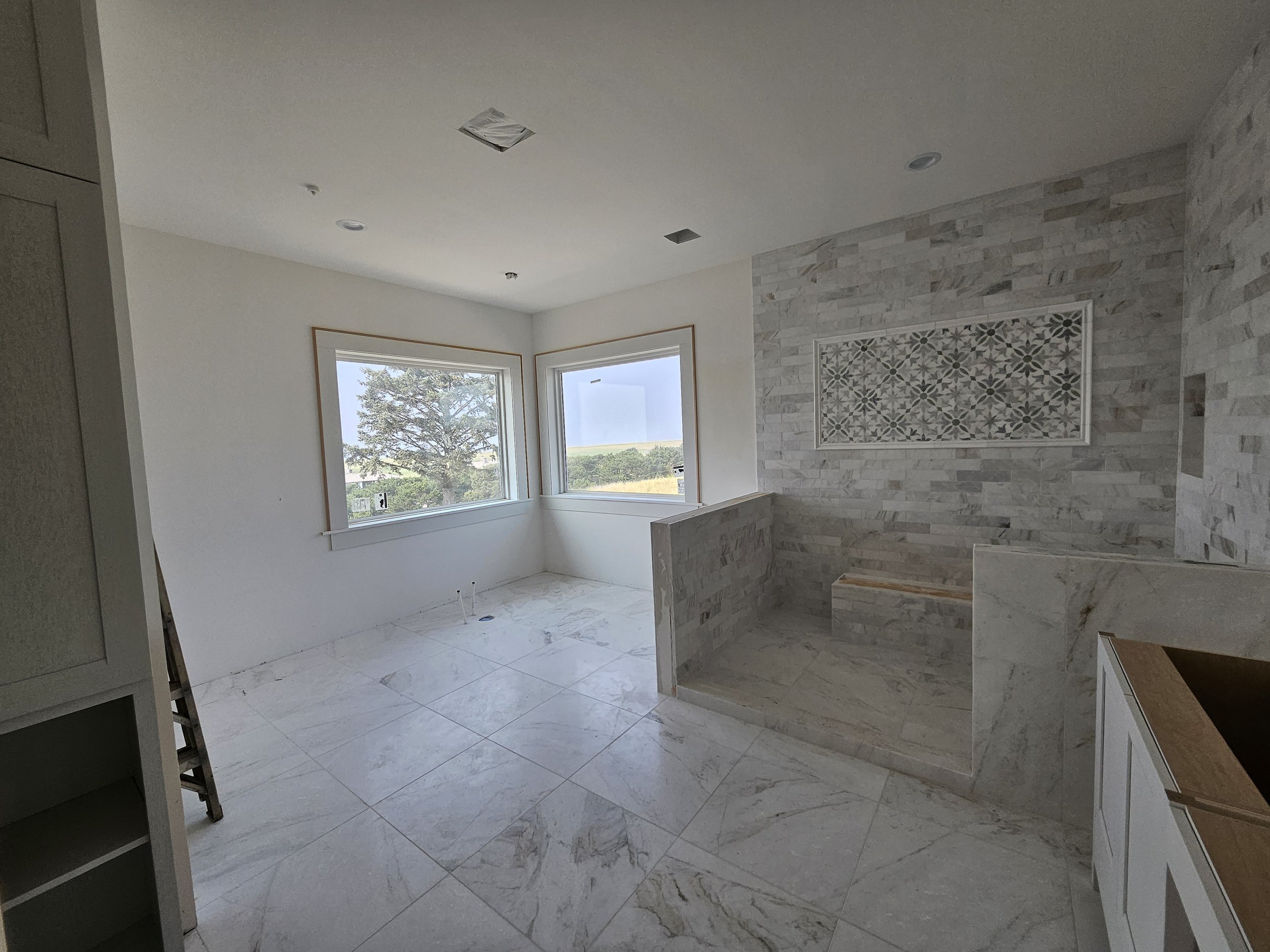 Interior of a room under construction or renovation with large windows, white walls, and a marble-like tiled floor and wall, featuring a decorative framed tile design.