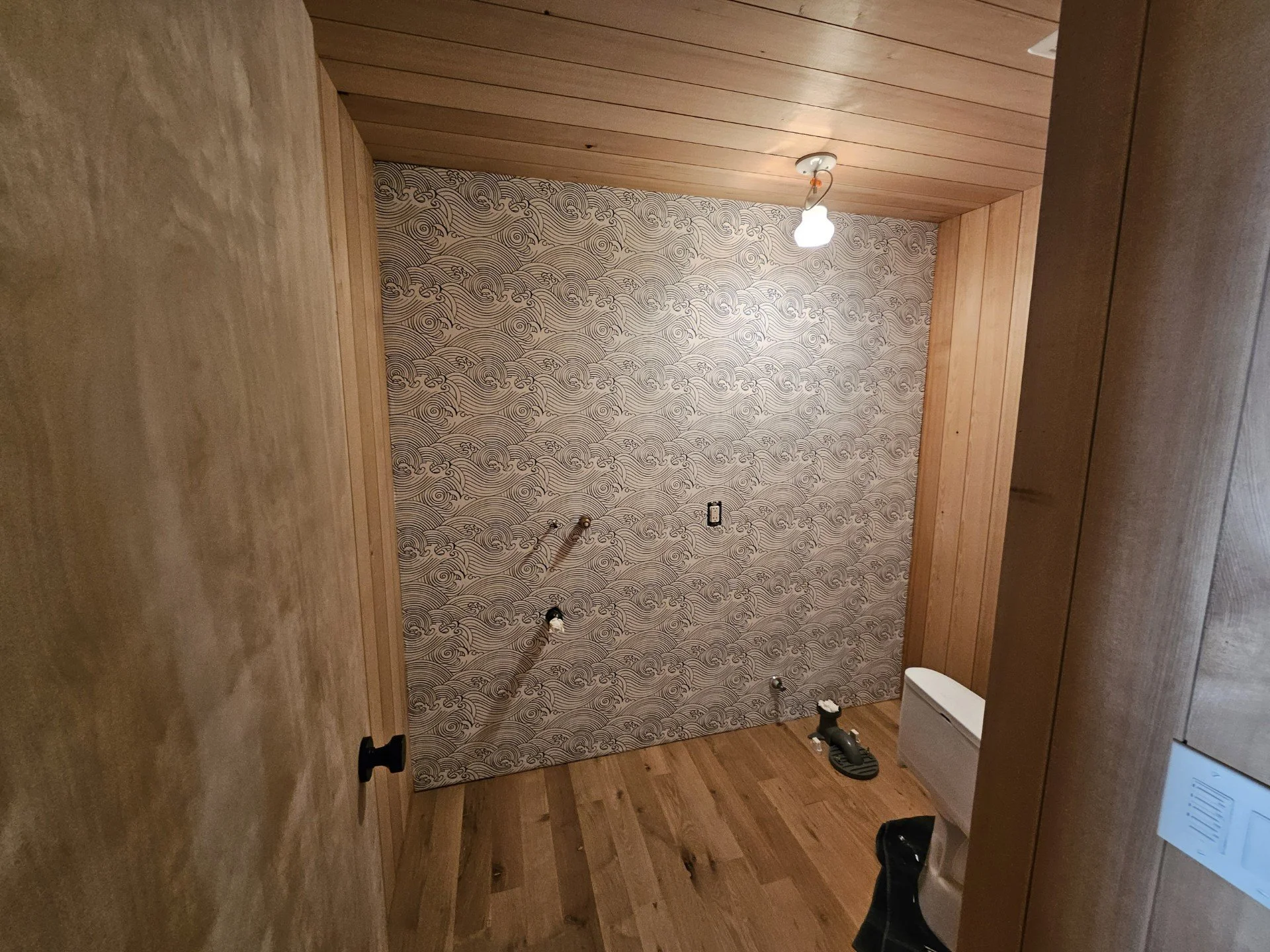 View of a small room under construction, with wallpapered wall featuring wave patterns, and wooden ceiling and floor. Construction tools and materials are visible on the floor.