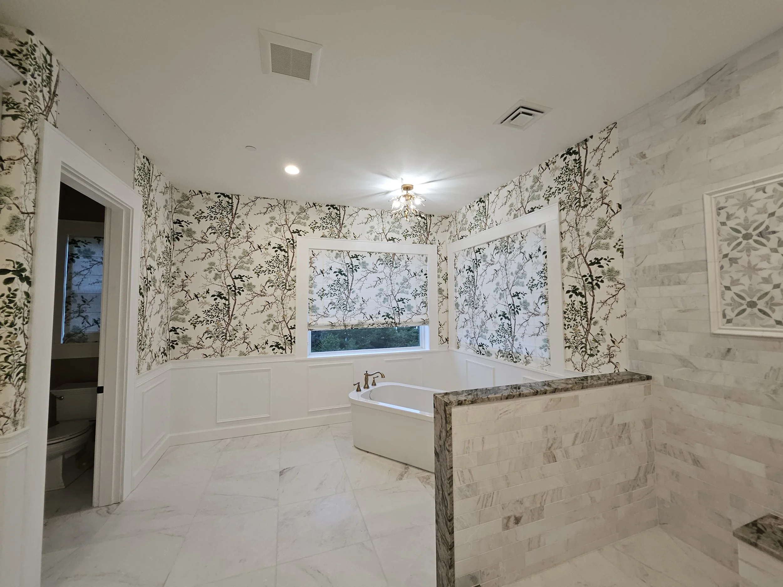 A bathroom with wallpaper featuring tree branches and leaves, a bathtub, a window showing green trees outside, and a partial view of a doorway to a toilet room.