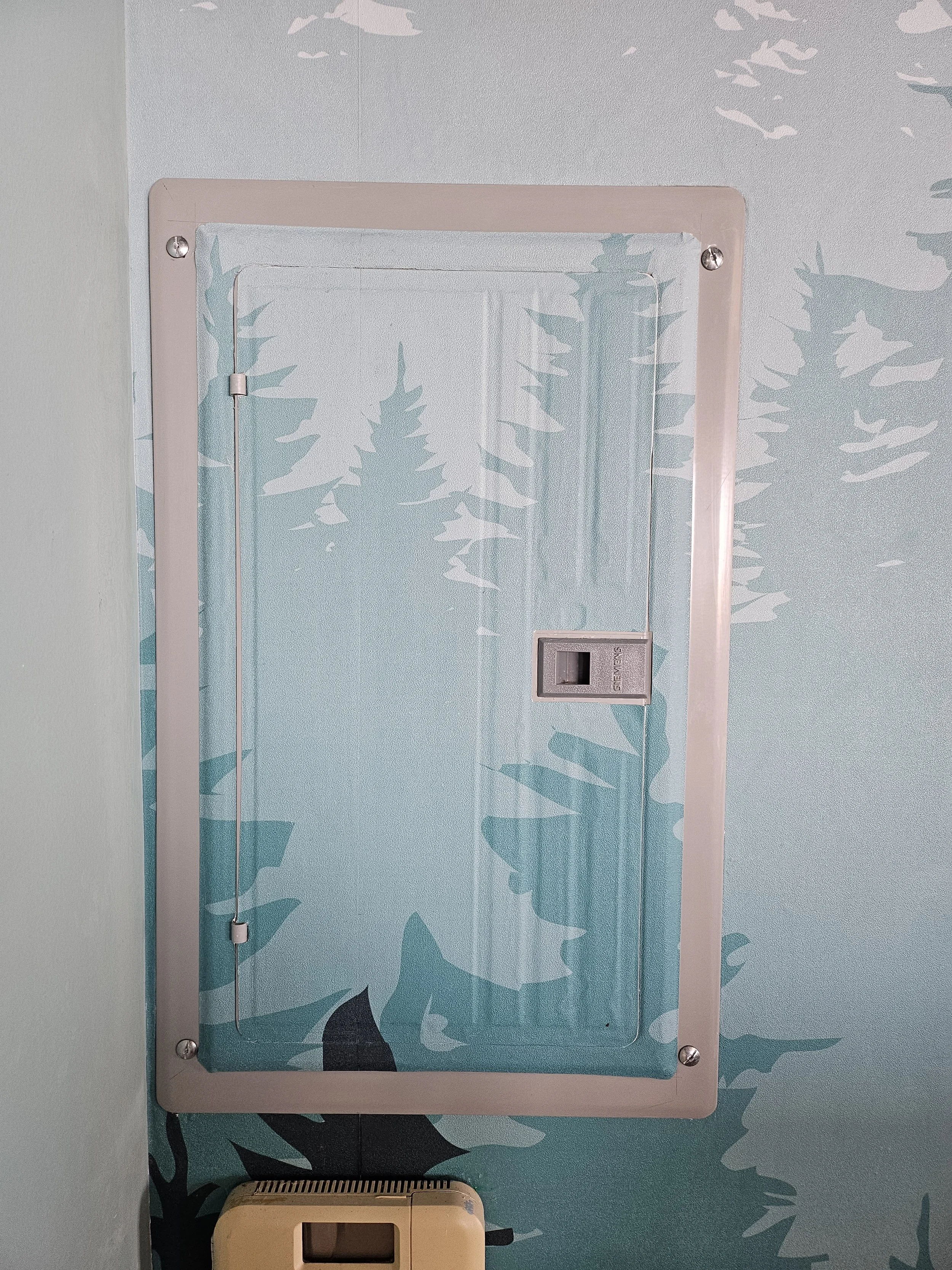 A wall-mounted mirror with a metal frame and four screws in each corner, reflecting a blue wall with a whale and ice floes mural. A beige box with a transparent front is underneath the mirror.