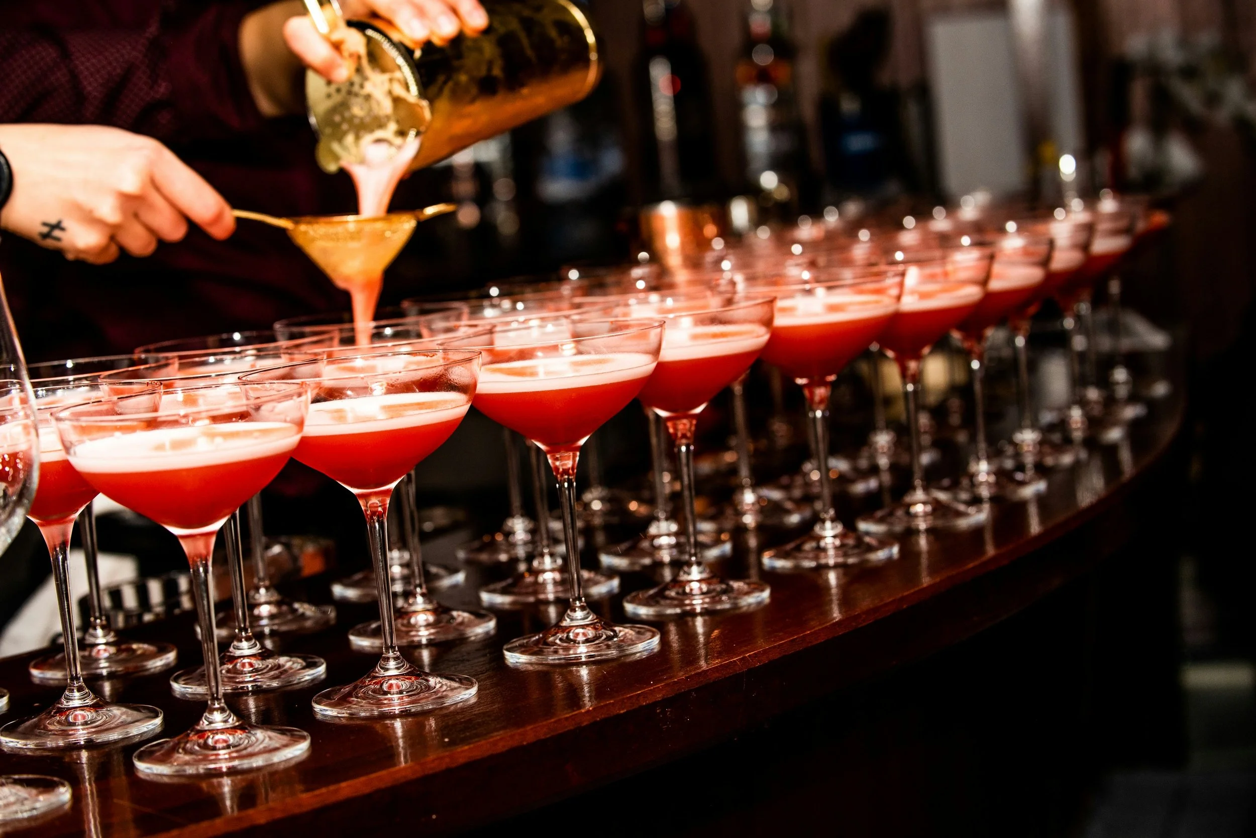 A bartender pours a pink cocktail from a shaker into a line of cocktail glasses on a wooden bar.