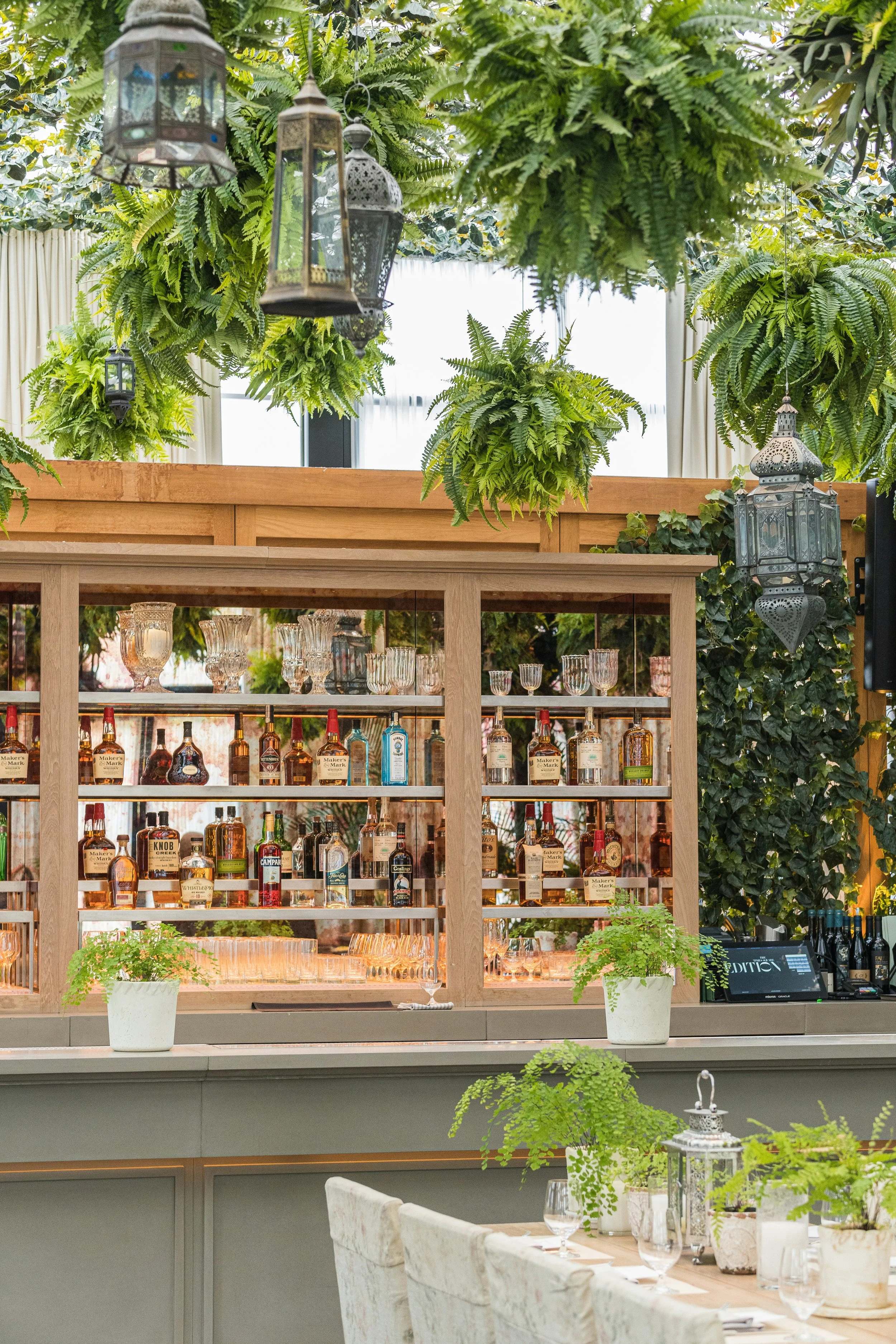 Interior of a restaurant or bar with a wooden display of liquor bottles and glassware, decorated with hanging green ferns and lanterns, and white draped curtains in the background.