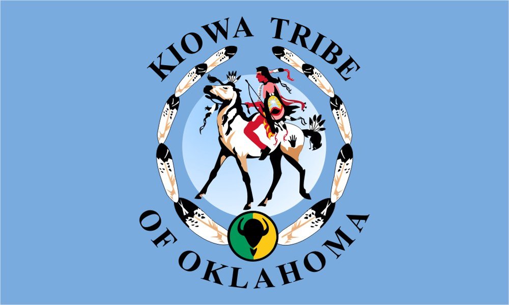 The flag of the Kiowa Tribe of Oklahoma with a blue background, featuring a central image of a warrior riding a horse, surrounded by black and white feathers, and a smaller circular emblem with a black outline, green background, and black image in the center, along with the text "Kiowa Tribe of Oklahoma" encircling the design.
